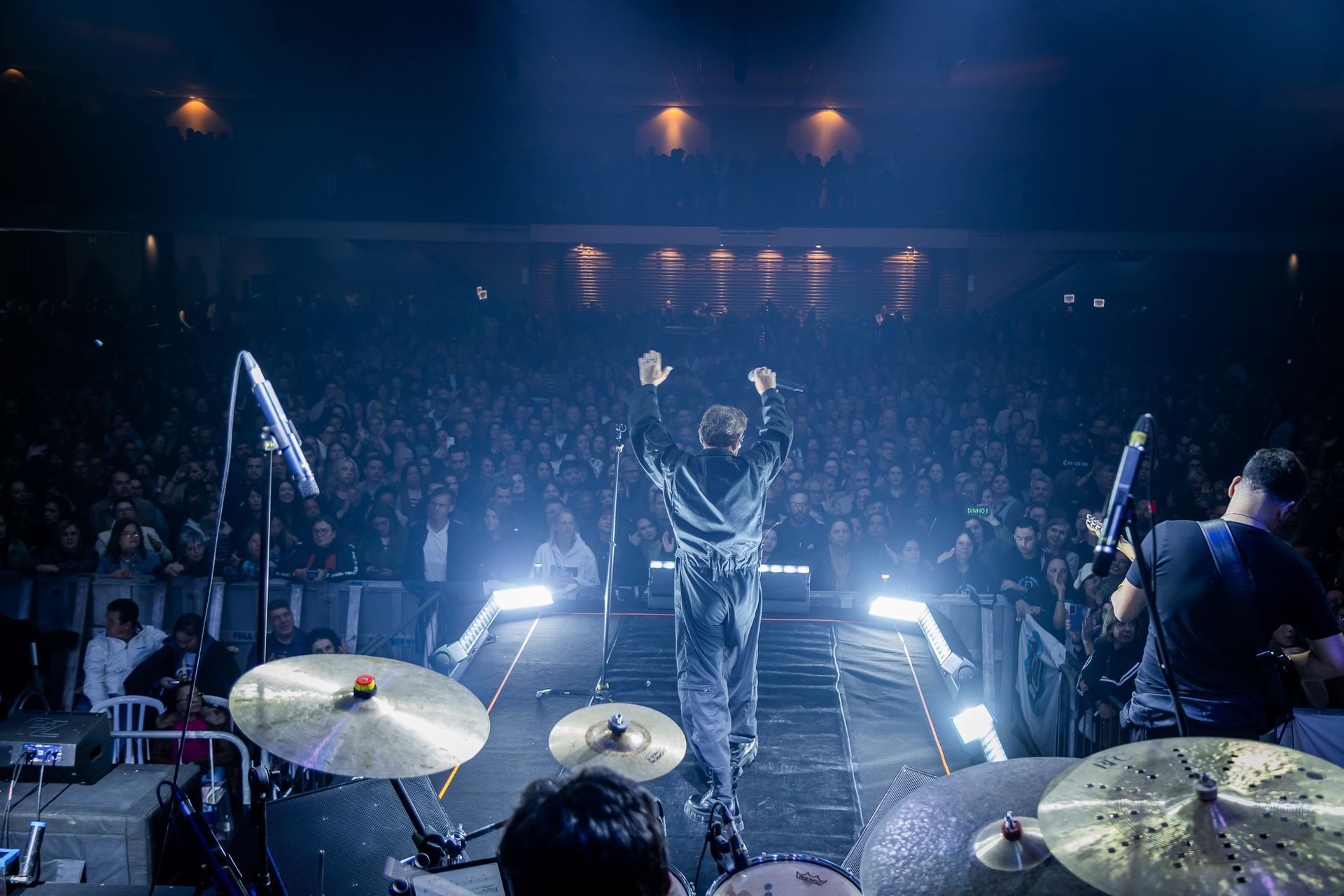 Um artista no palco com os braços erguidos, de frente para uma grande plateia iluminada em um show.