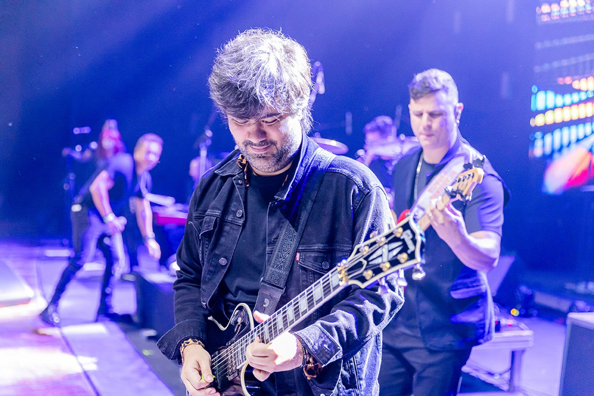 Uma banda de rock se apresenta no palco sob luzes azuis, com um guitarrista em primeiro plano e um baixista atrás dele.