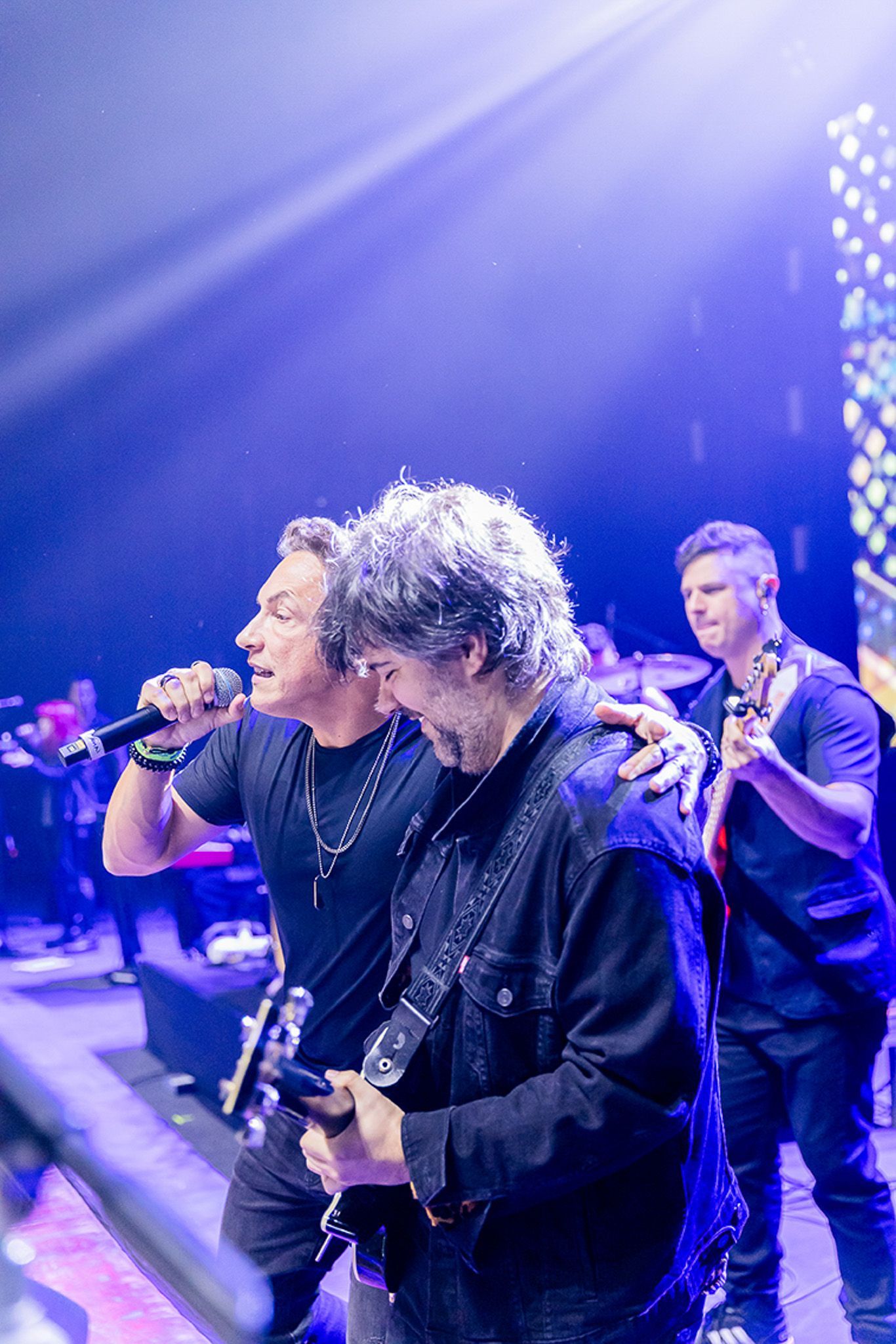 Três músicos se apresentam em um palco iluminado por uma luz azul brilhante, tocando guitarras e cantando em um microfone.