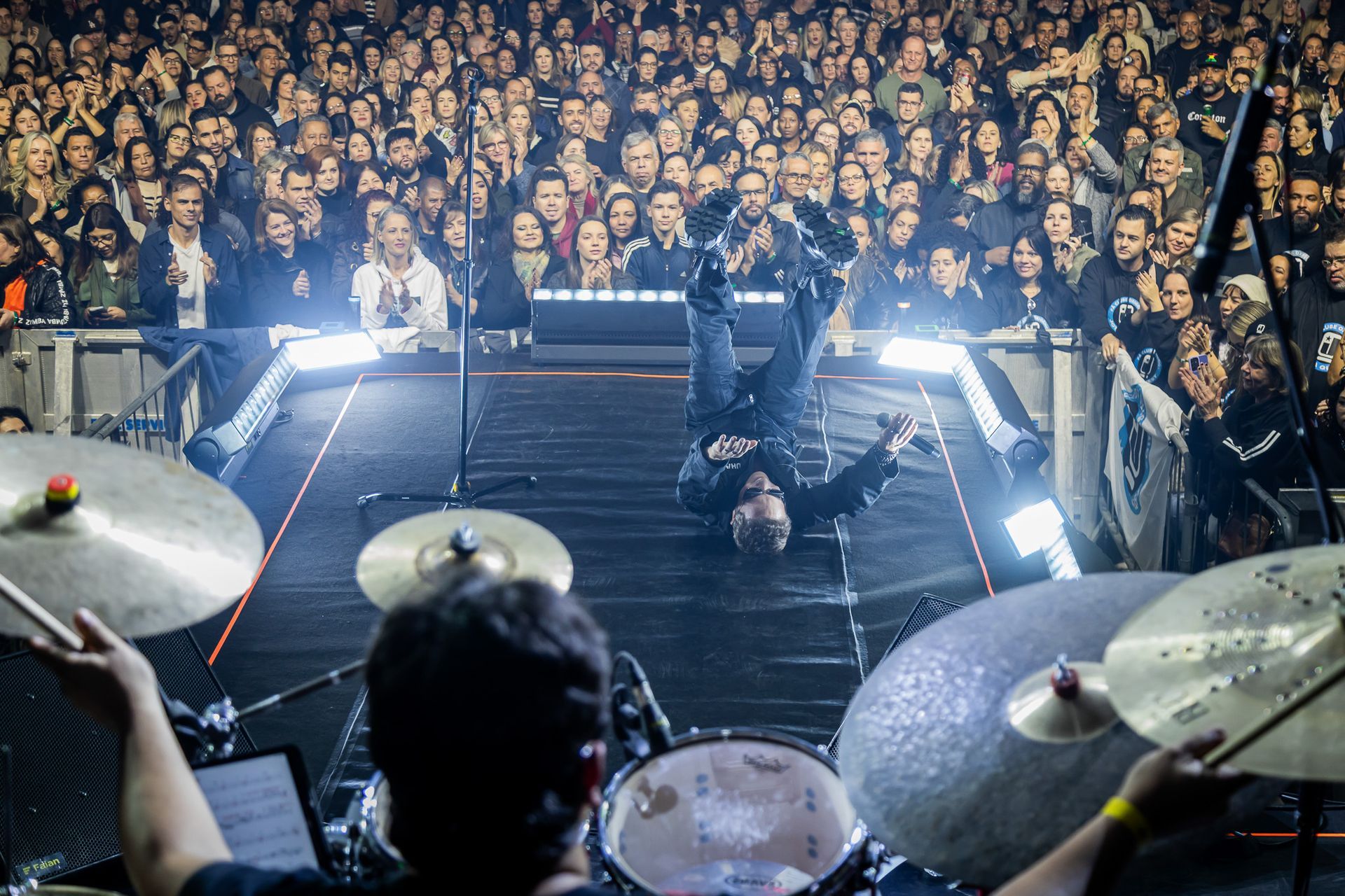 Um artista realiza uma parada de mãos no palco em frente a uma plateia, visto da perspectiva de um baterista.