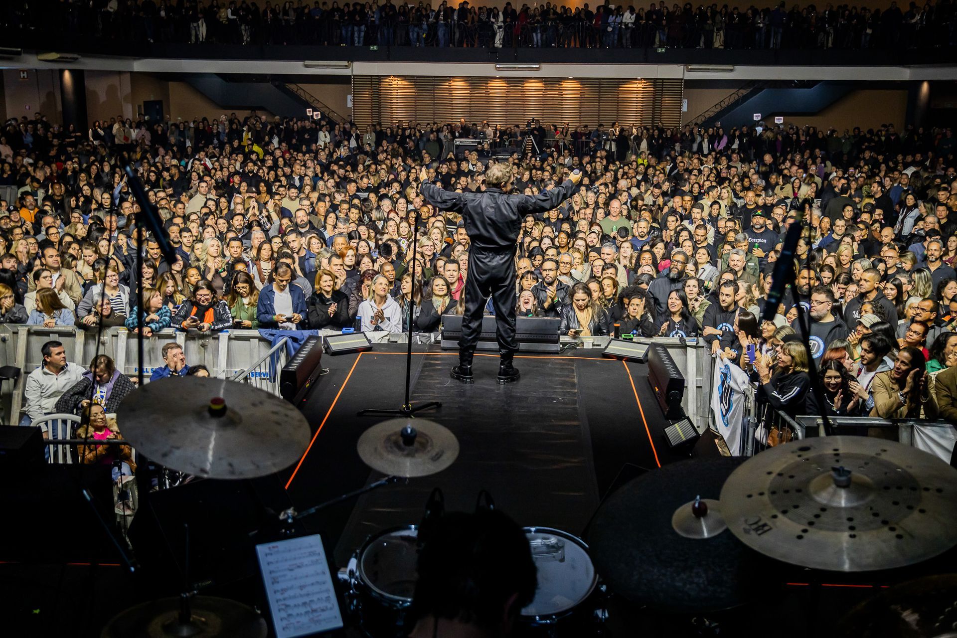 Um artista está no palco com os braços estendidos, de frente para uma grande e densa plateia em uma casa de shows fechada.
