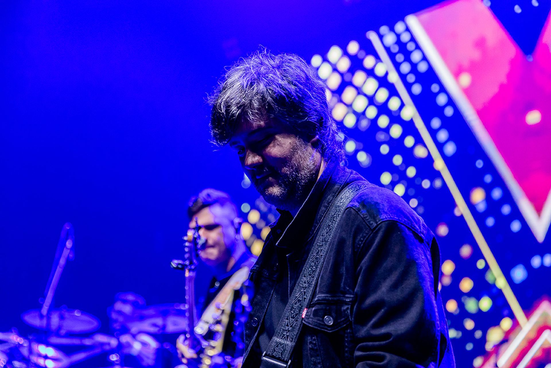 Um músico de cabelos ondulados se apresenta no palco vestindo uma jaqueta escura, com um colega de banda visível ao fundo.