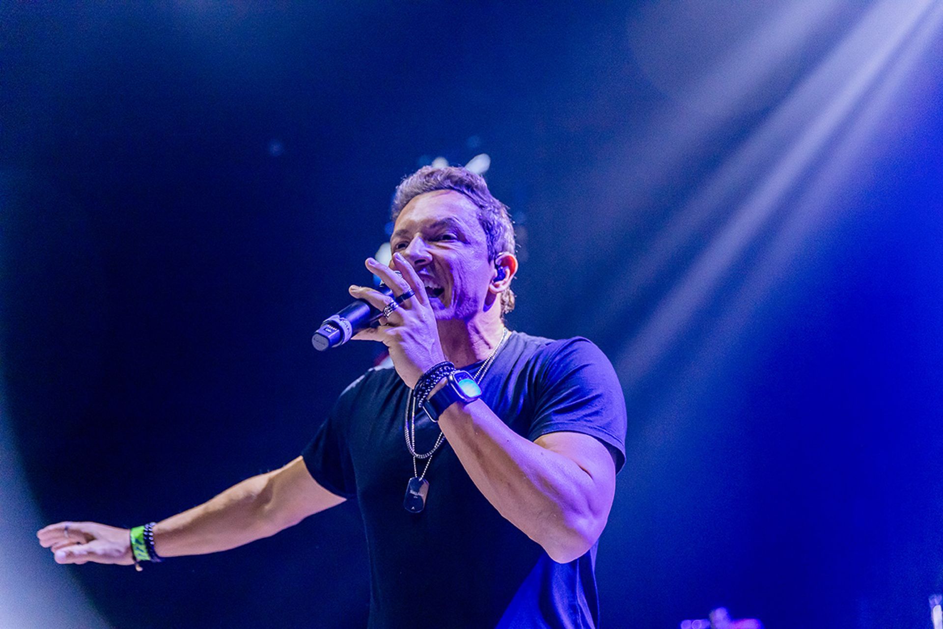 Um homem cantando em um microfone em um palco sob holofotes azuis, gesticulando com a mão em direção à plateia.