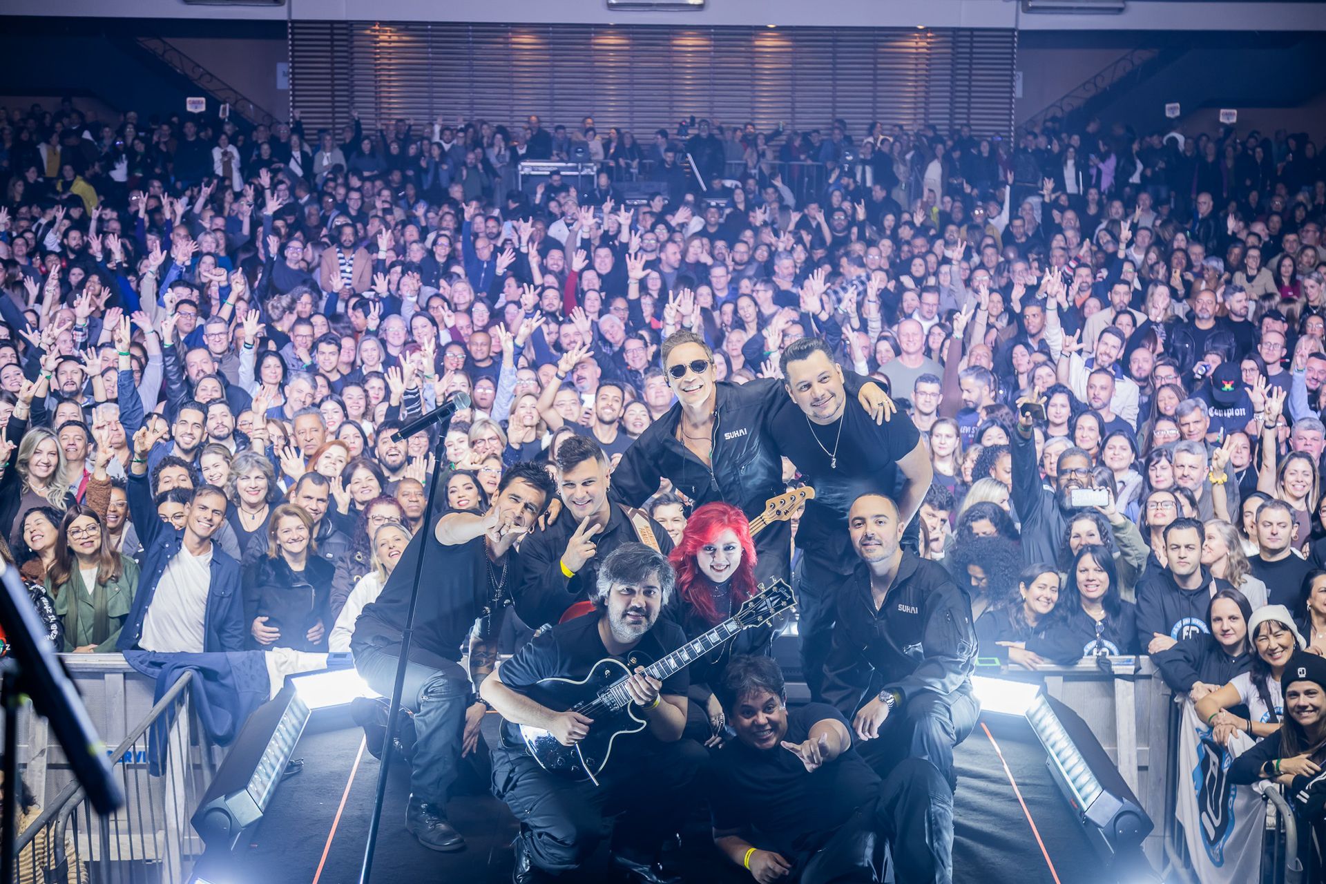 Uma banda posa para uma foto no palco com seus instrumentos em frente a uma grande plateia animada em um show em uma arena.
