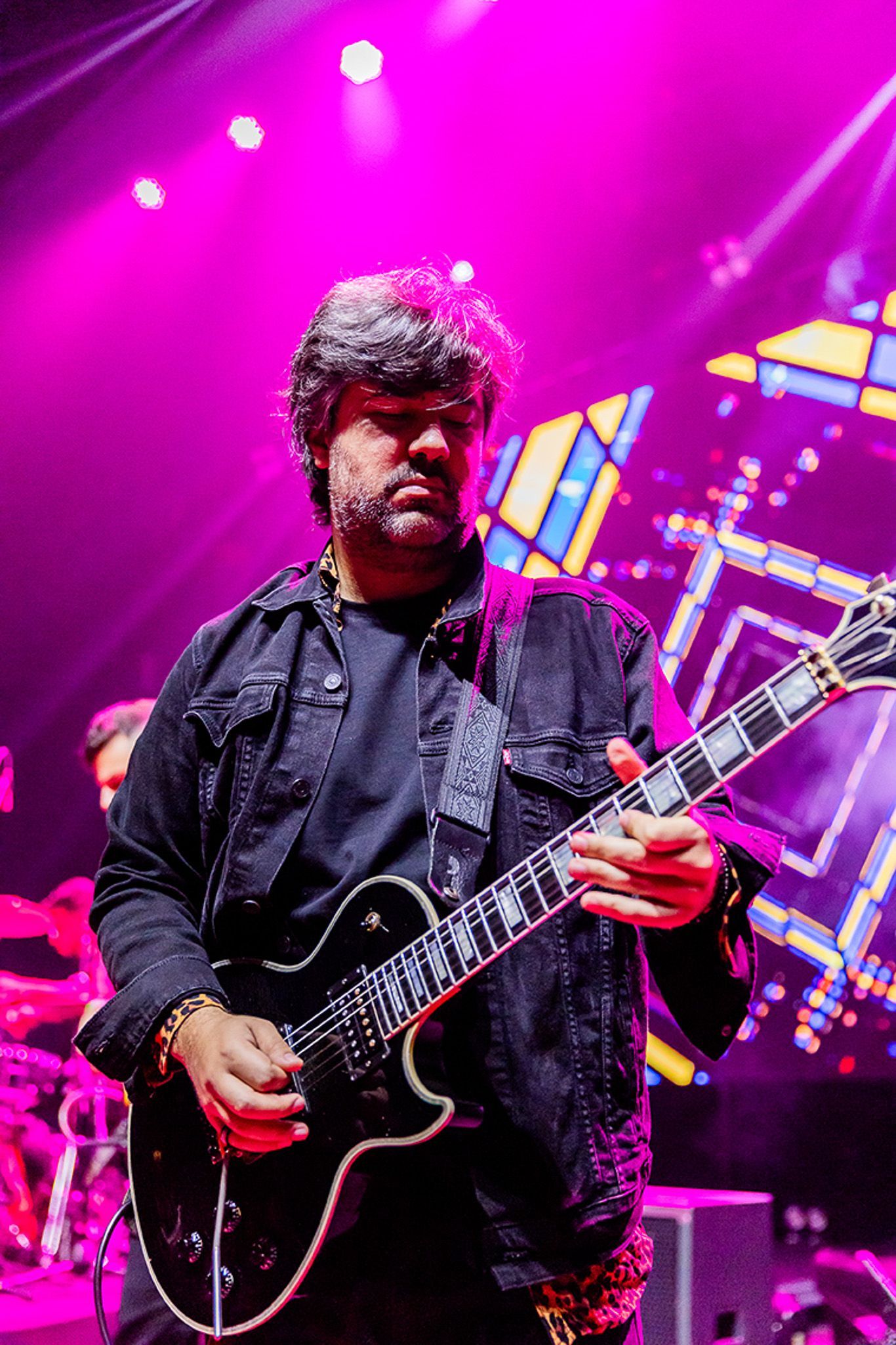 Um artista toca guitarra elétrica em um palco iluminado com luzes rosa vibrantes e um padrão geométrico de fundo.