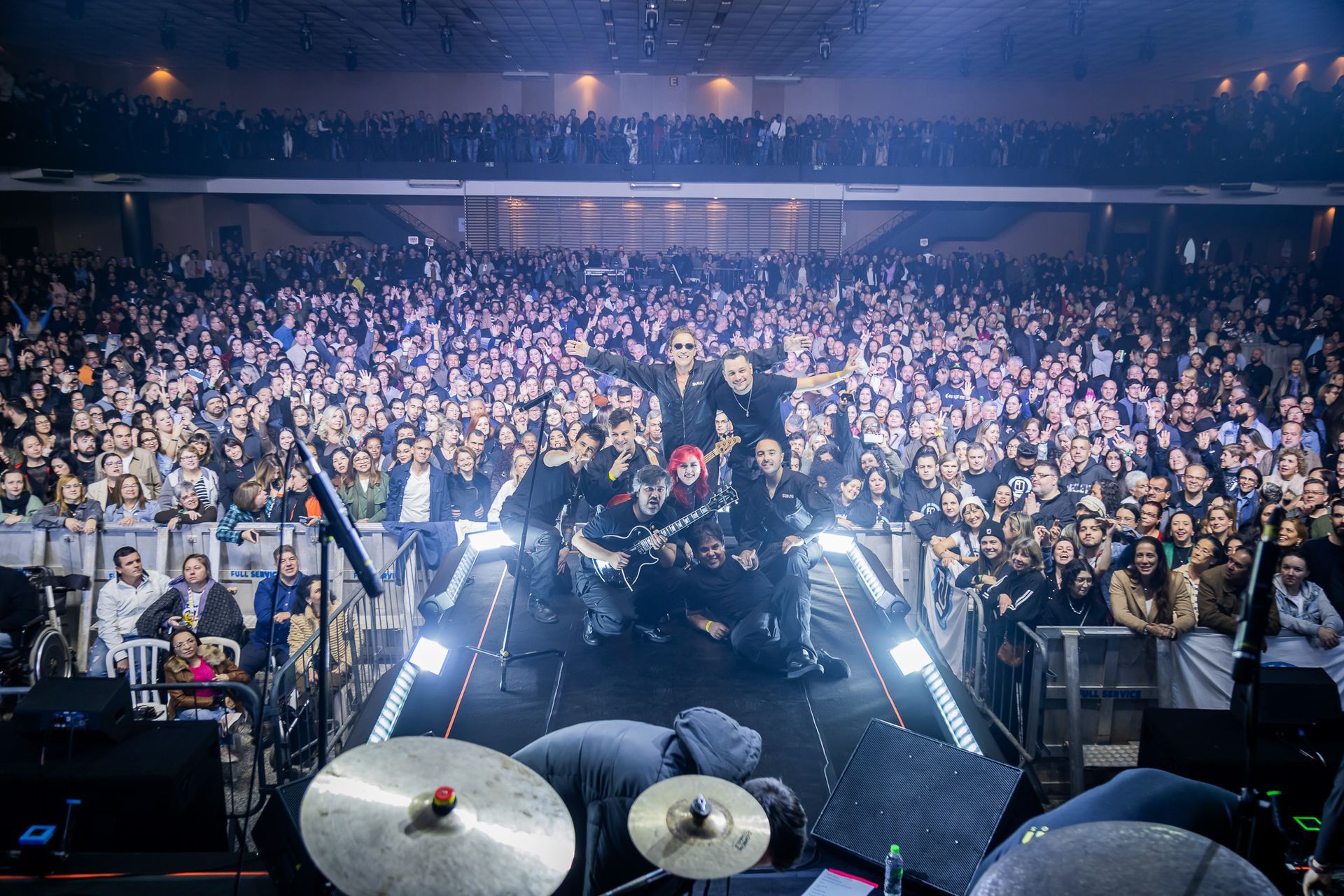 Uma banda se apresentando no palco em uma casa de shows lotada, com os músicos posando diante de uma grande plateia.