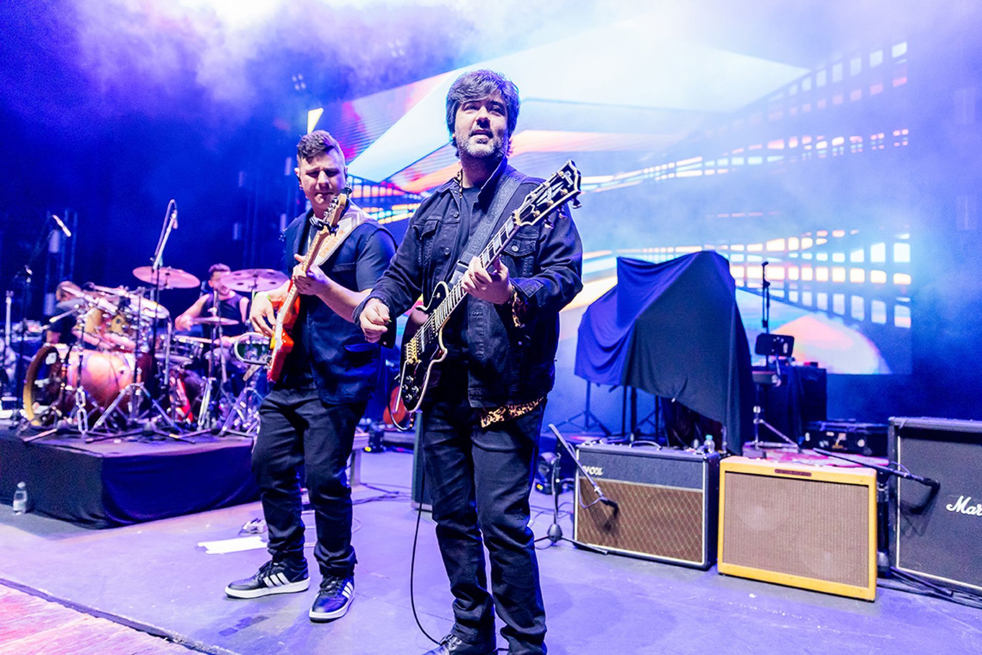 Dois guitarristas se apresentam em um palco com um baterista ao fundo, cercados por luzes de palco azuis e equipamentos.