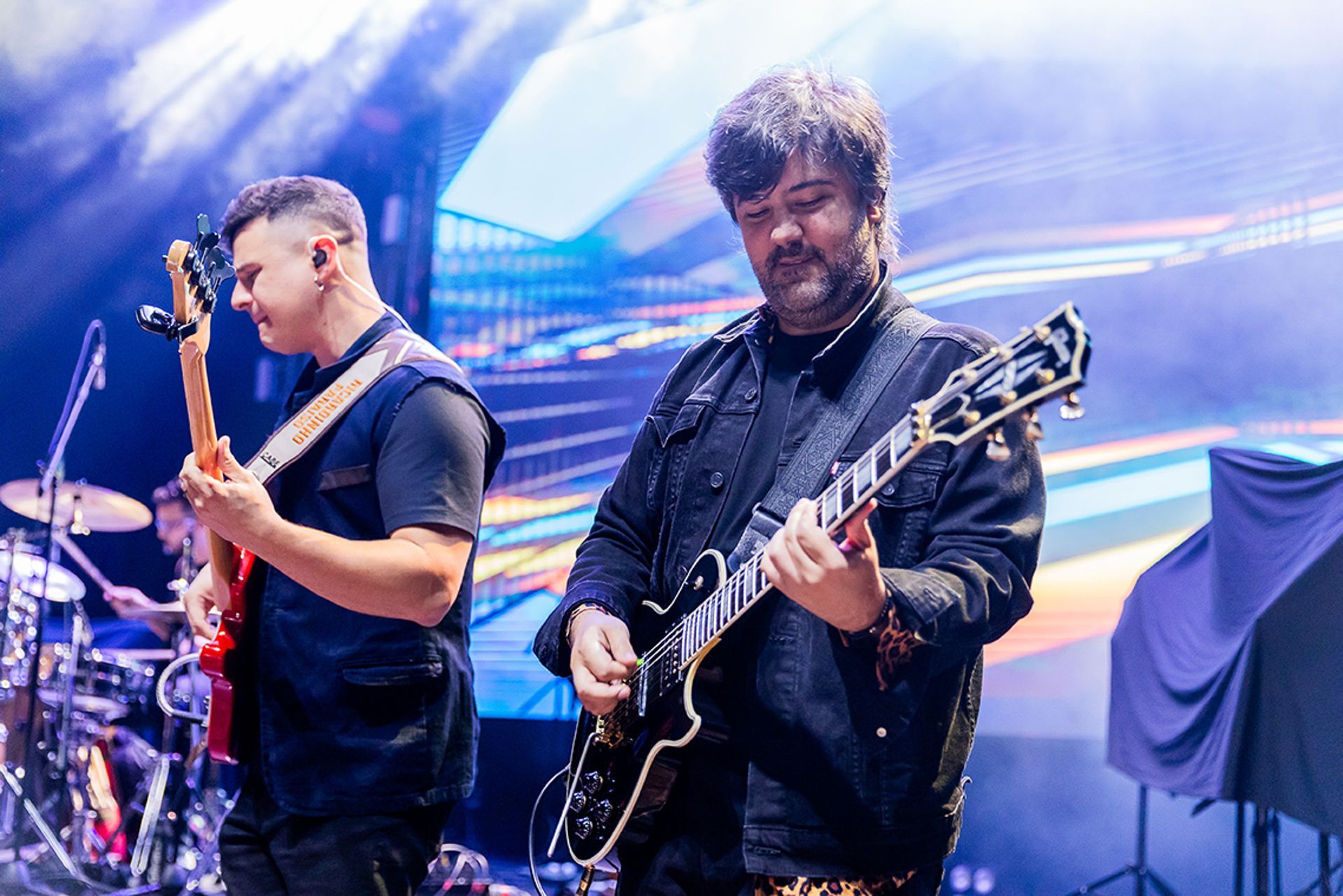 Dois músicos se apresentam no palco; um toca um baixo elétrico vermelho e o outro toca uma guitarra elétrica preta.