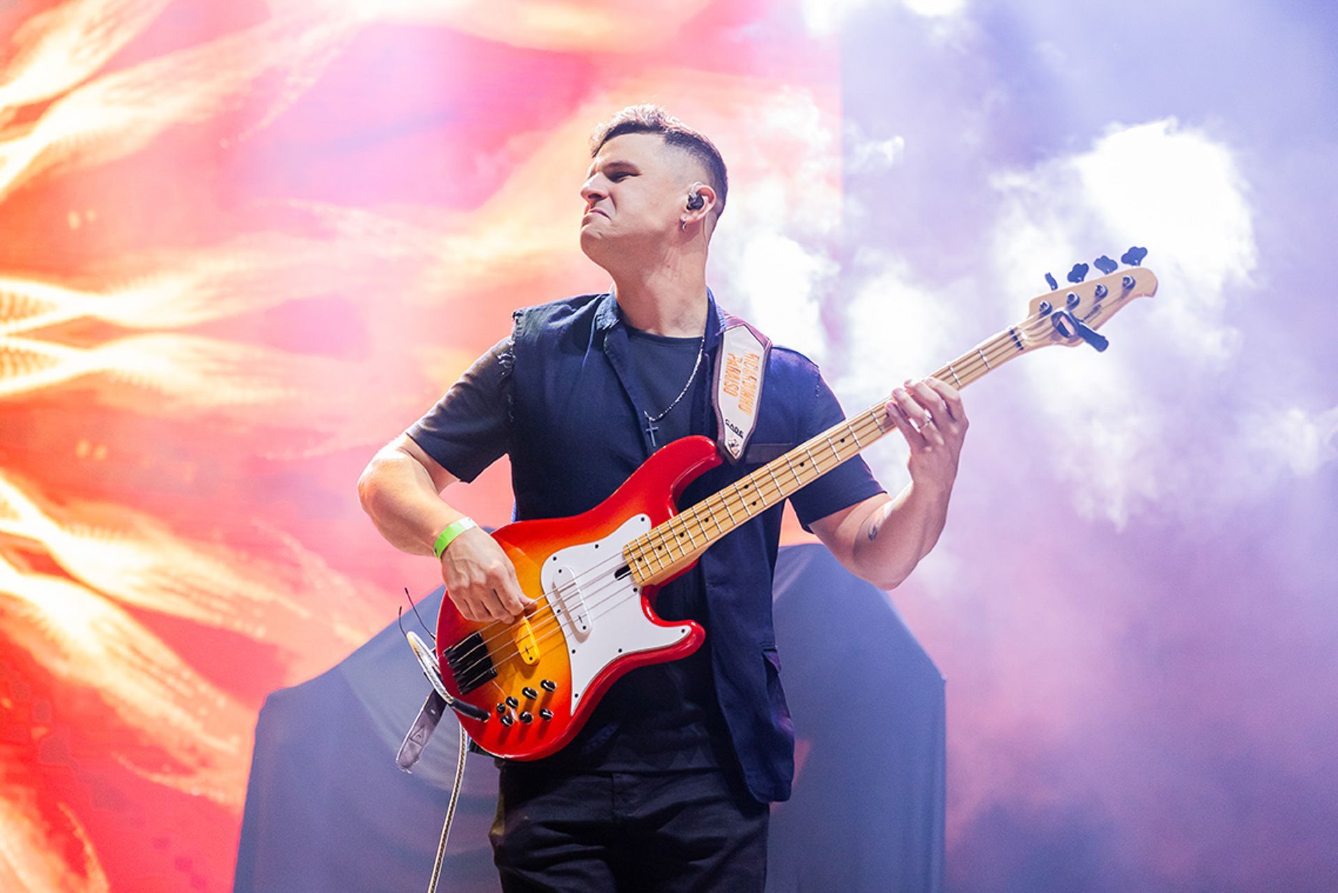 Um músico tocando um baixo elétrico vermelho e laranja em um palco com um fundo colorido e vibrante.