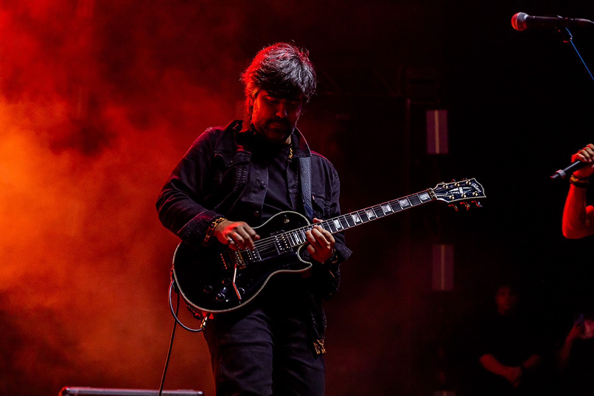 Um músico toca uma guitarra elétrica preta no palco, iluminado por uma dramática luz vermelha e fumaça.