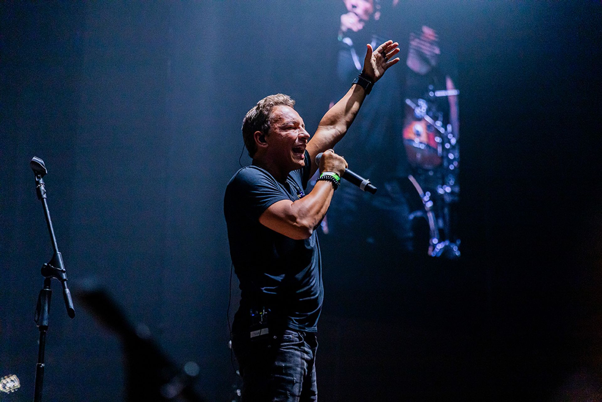 Um cantor com um microfone está no palco sob holofotes azuis, erguendo um braço em direção à plateia.