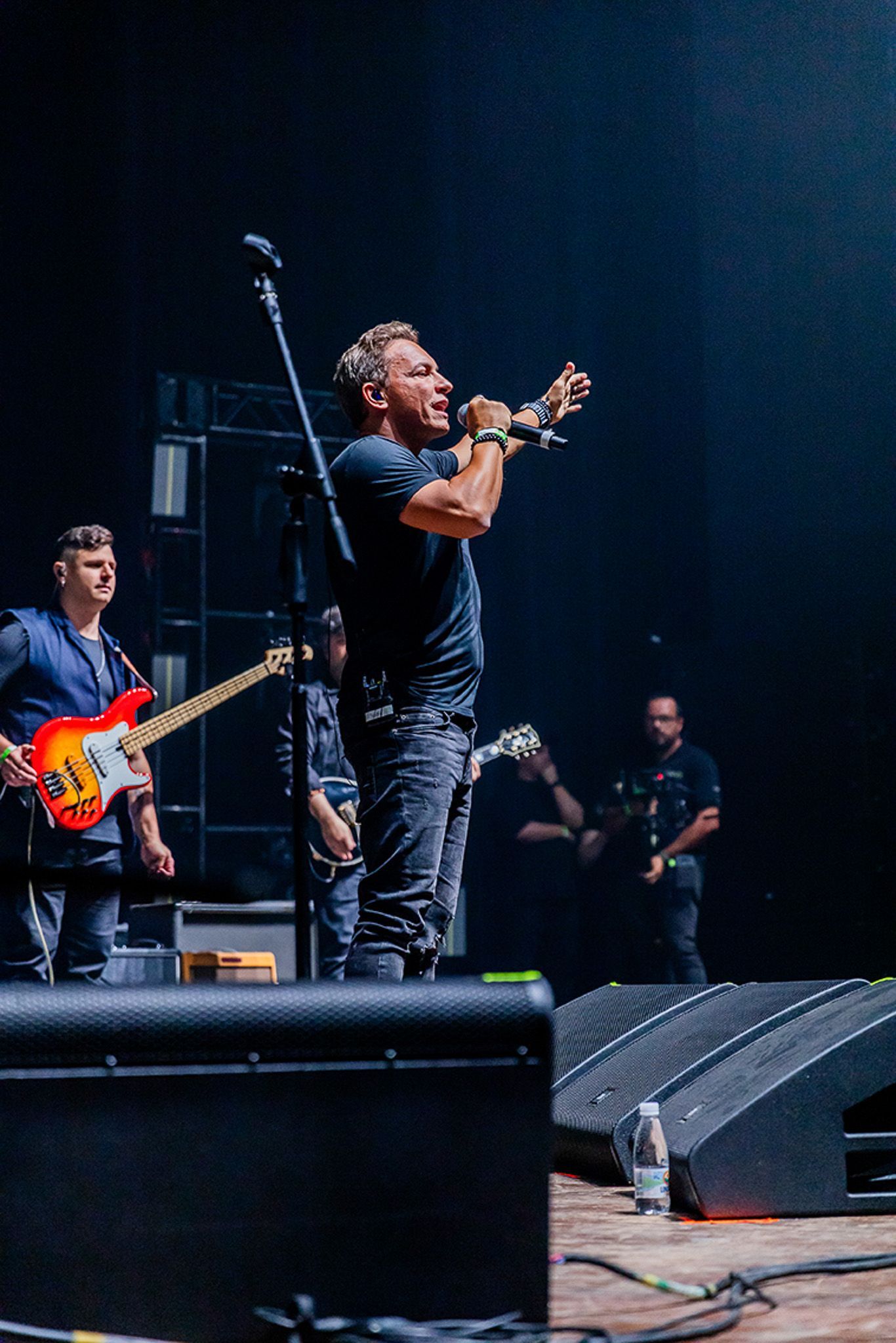 Um artista cantando no palco com um microfone, acompanhado por um baixista e outros membros da banda.