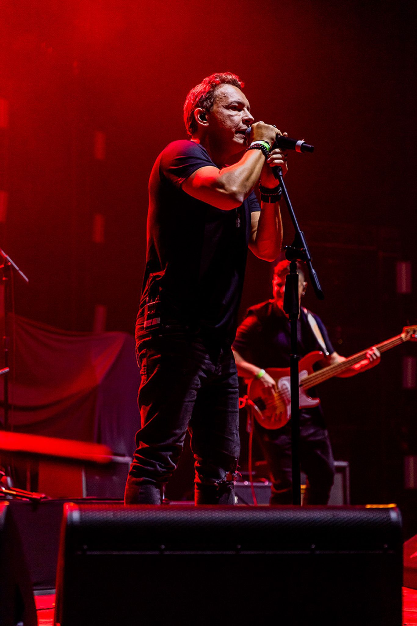 Um cantor se apresenta em um microfone em um palco com iluminação, enquanto um baixista toca ao fundo.