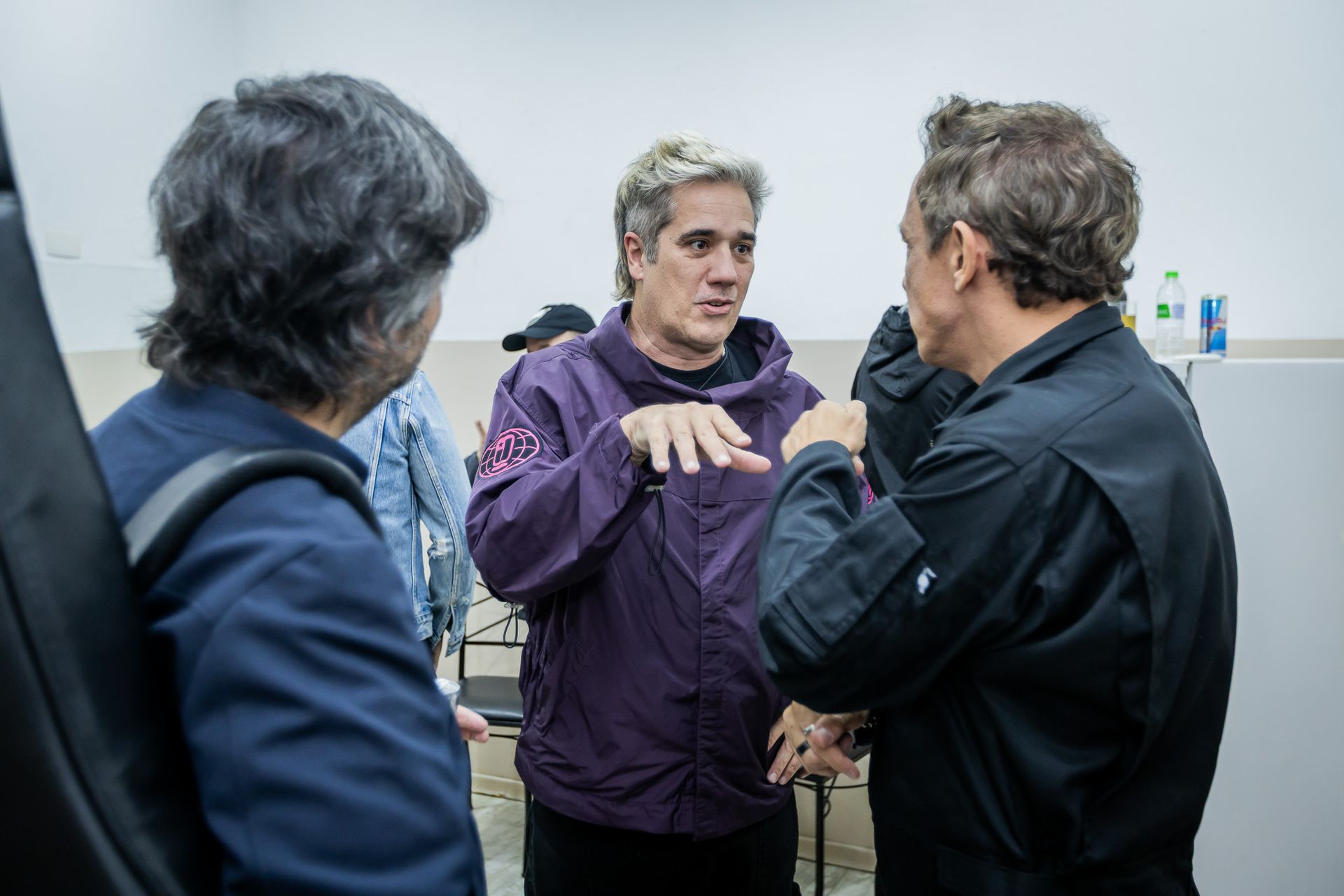 Três pessoas conversando em uma sala bem iluminada. A pessoa no meio veste uma jaqueta roxa e gesticula enquanto fala.