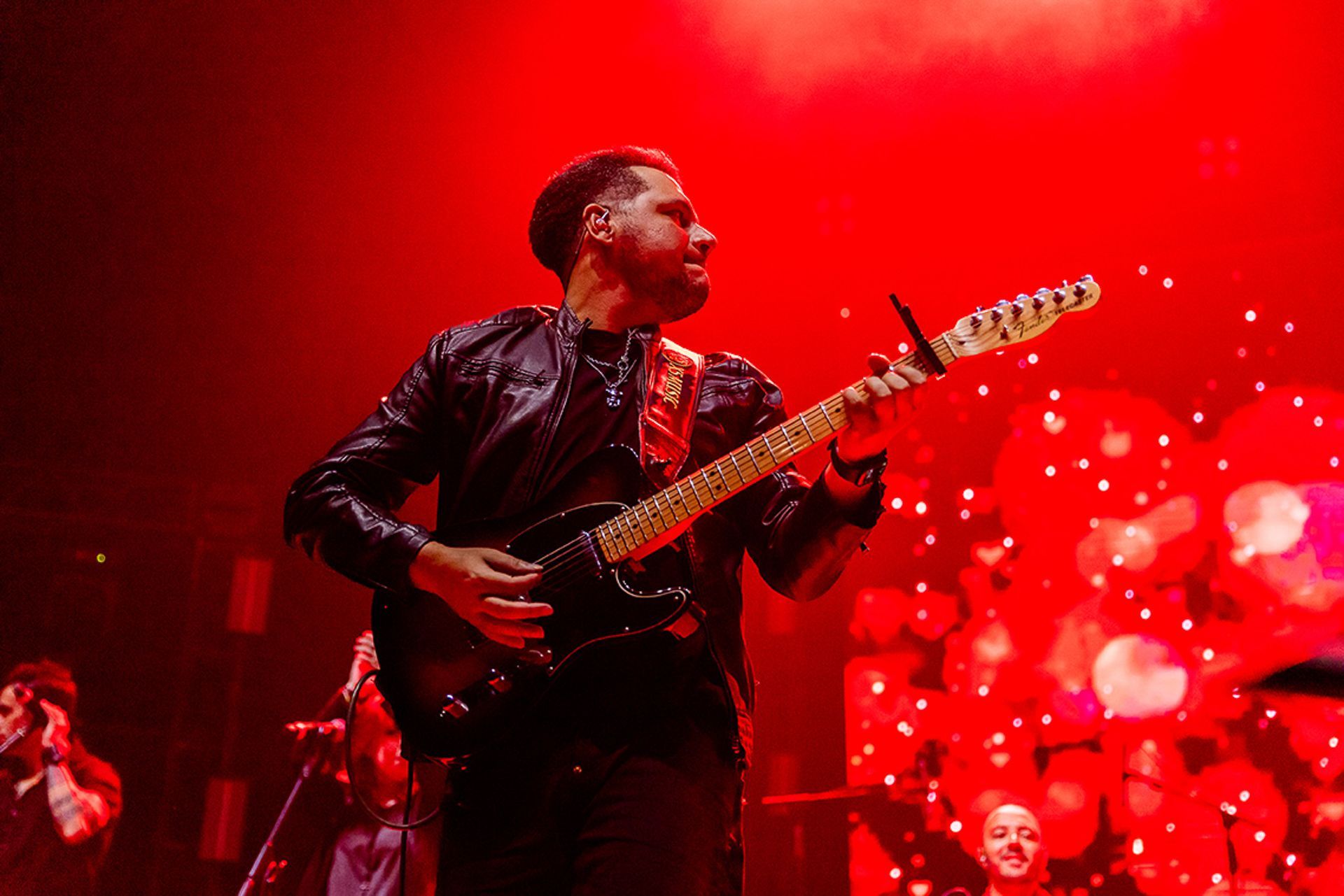 Um músico toca uma guitarra elétrica preta no palco sob uma iluminação vermelha vibrante, com uma multidão visível ao fundo.