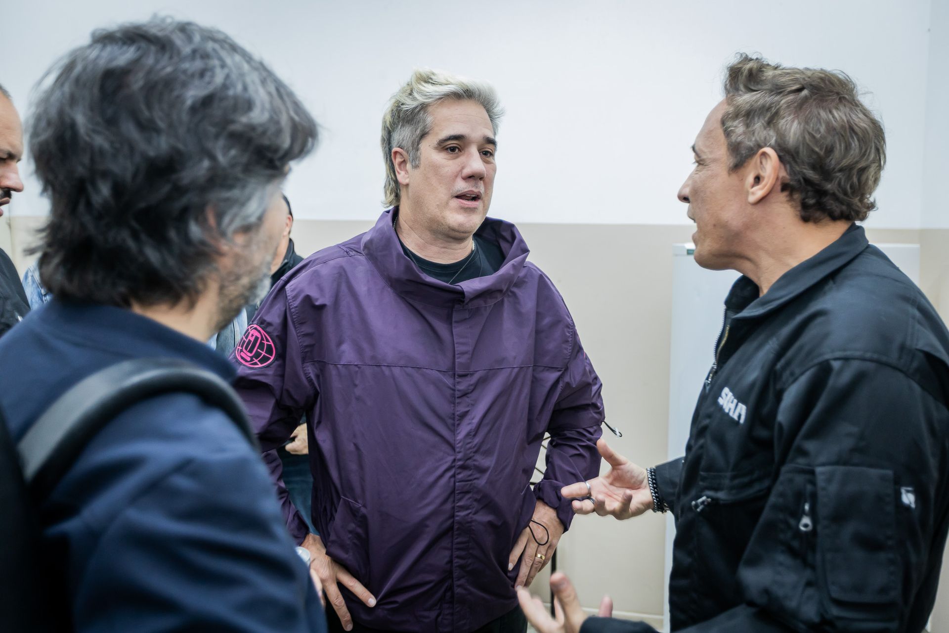 Três indivíduos de jaqueta participam de uma discussão séria e animada em um espaço interno com paredes lisas.