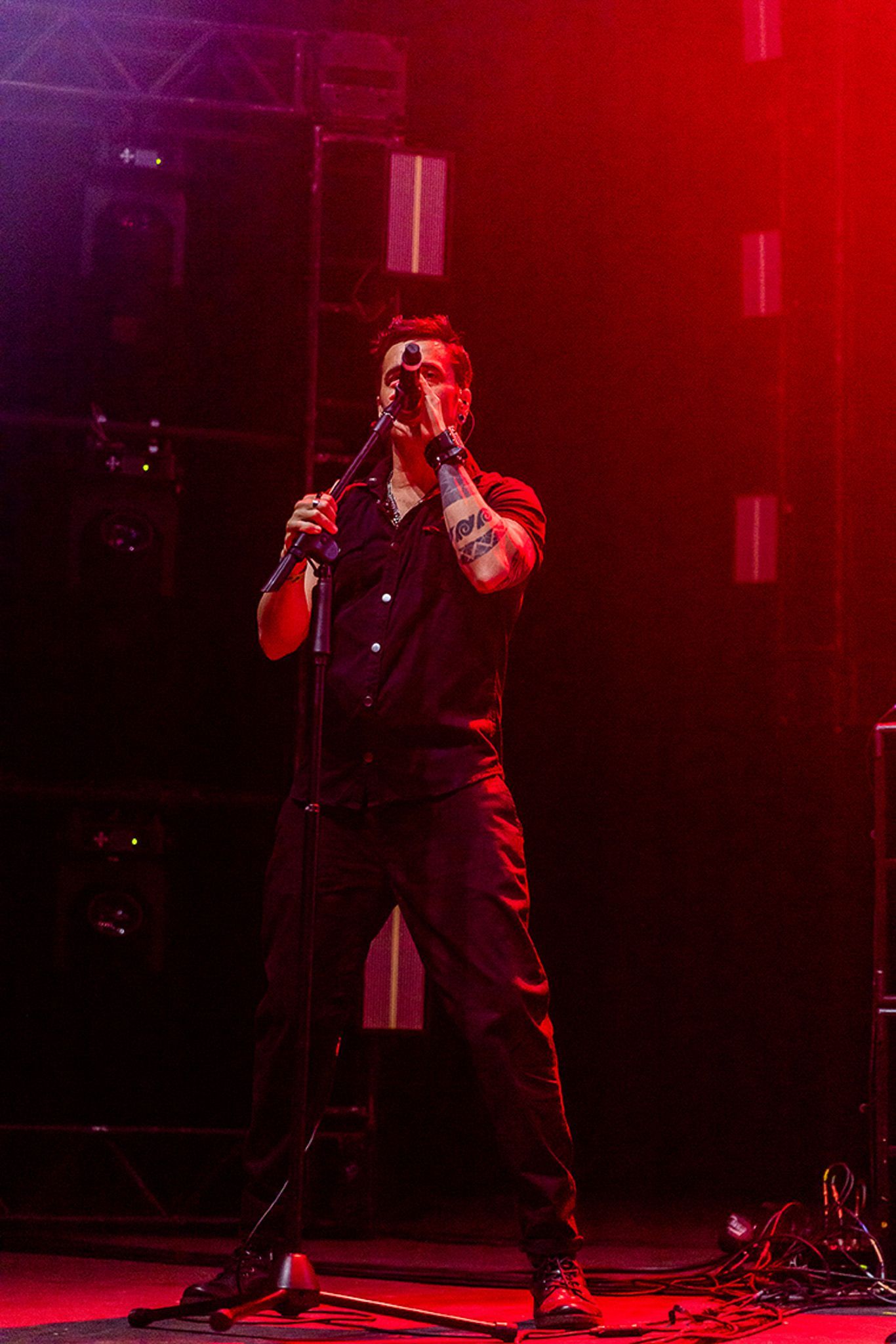 Um artista cantando em um microfone em um palco iluminado com luz vermelha, vestindo uma camisa escura de botões e calças.