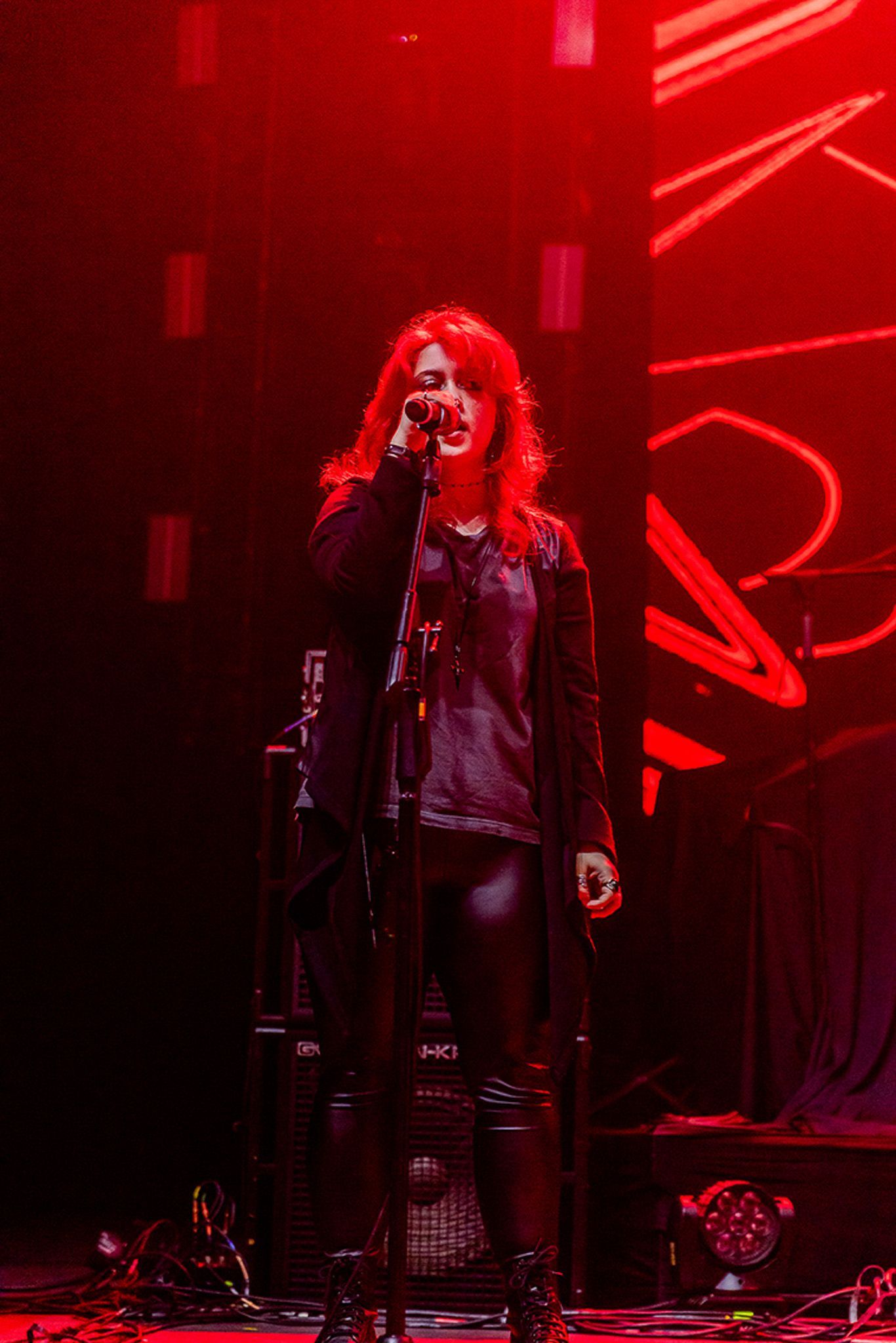 Uma artista de longos cabelos ruivos canta em um microfone no palco, banhada por uma vibrante iluminação vermelha.