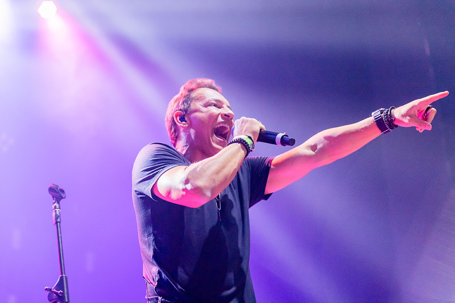 Um cantor se apresenta no palco sob luzes roxas, cantando em um microfone com um braço estendido e apontando para a frente.