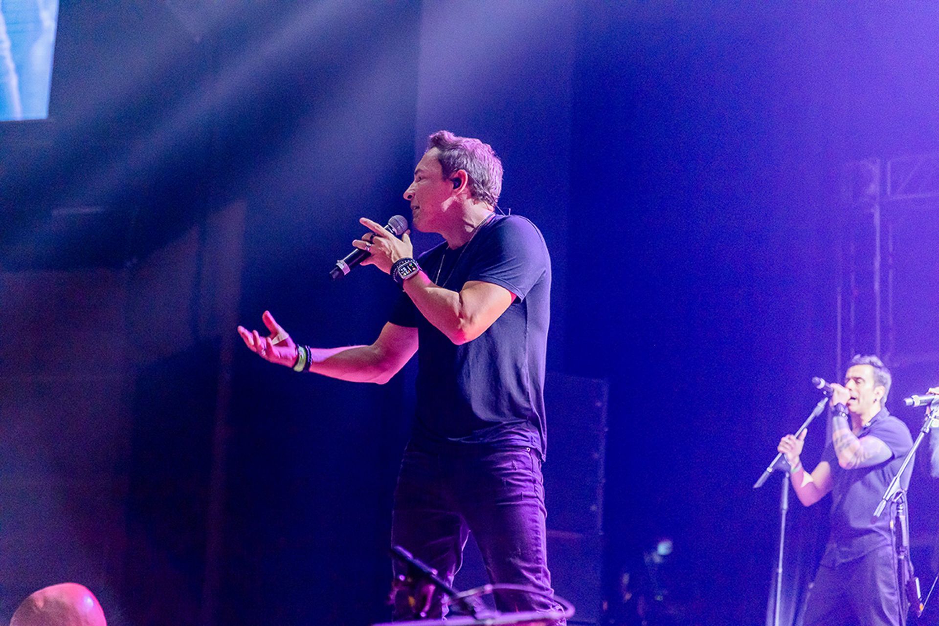 Um artista canta em um palco com iluminação roxa vibrante, gesticulando para a plateia.