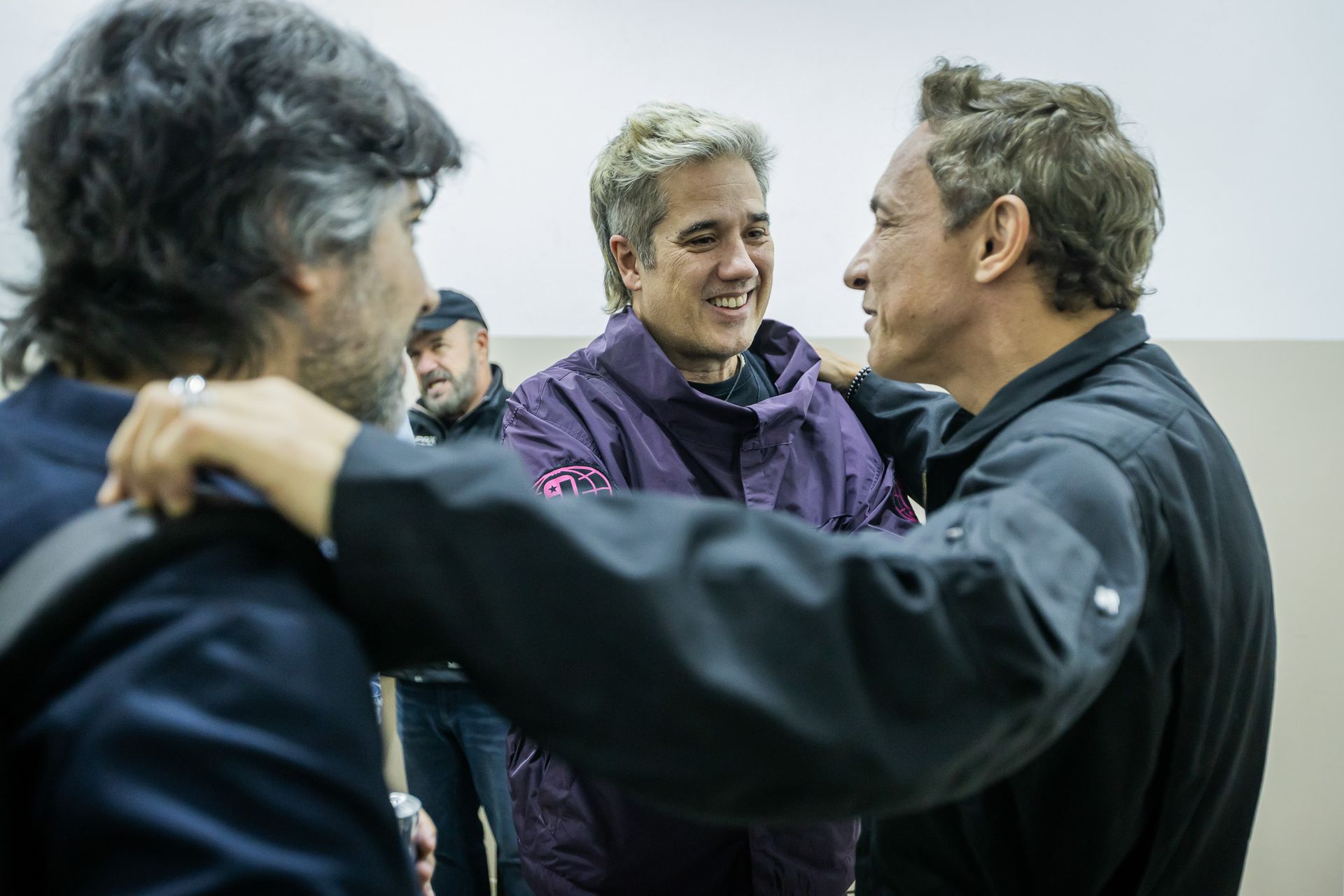 Três pessoas vestidas casualmente interagem em um ambiente interno simples; uma sorri enquanto as outras observam.