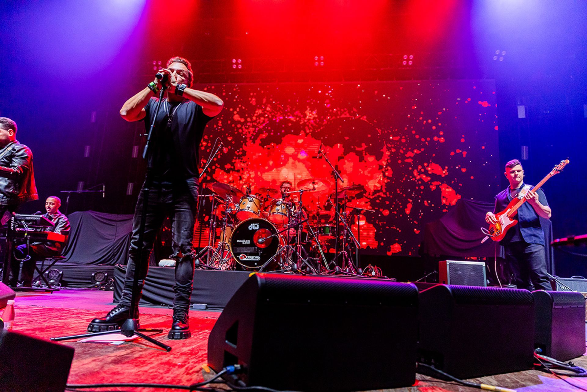 Uma banda se apresenta em um palco iluminado, composta por um vocalista, um guitarrista e um baterista.