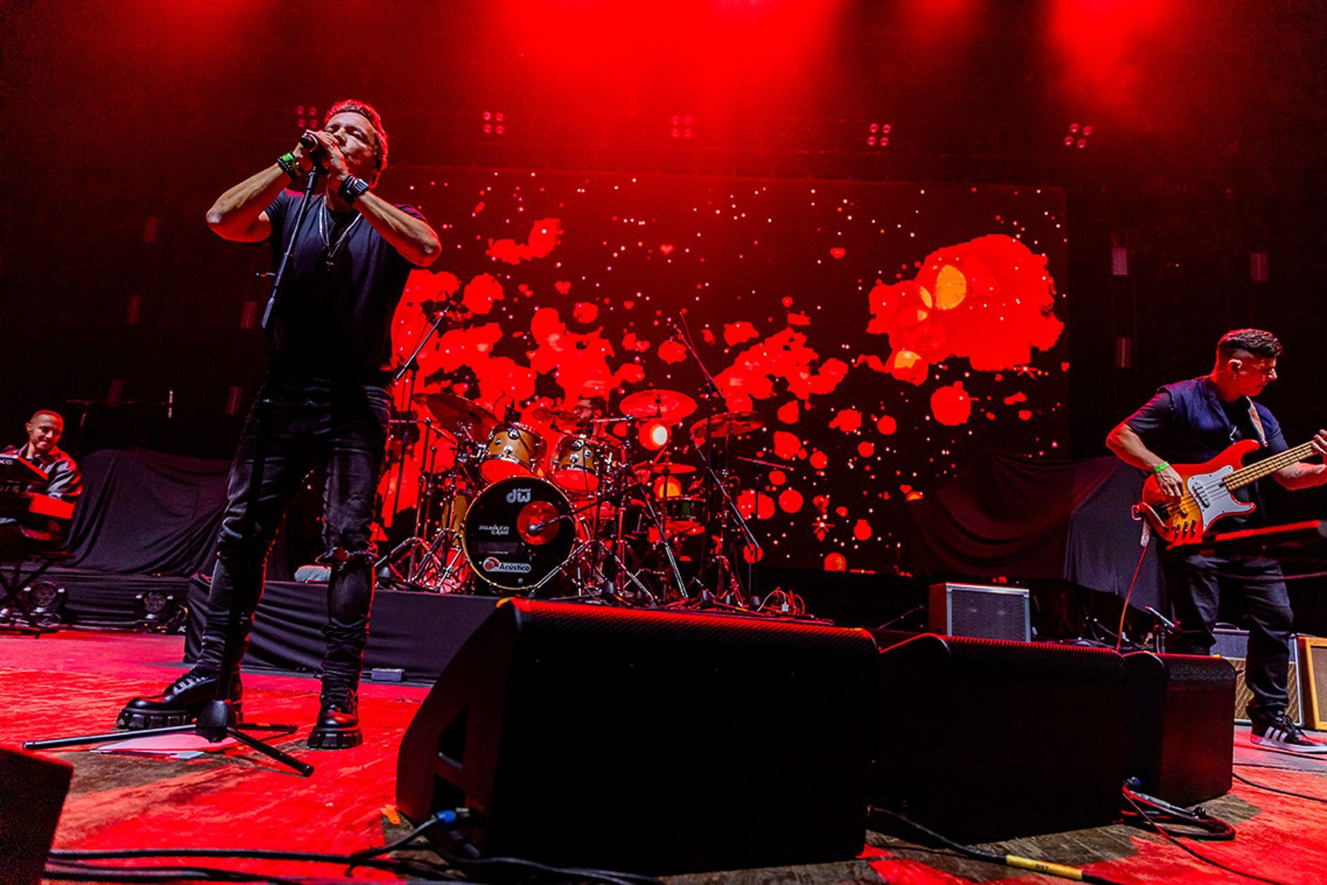 Uma banda de rock se apresenta no palco sob luzes vermelhas brilhantes, com um vocalista e um baixista tocando.