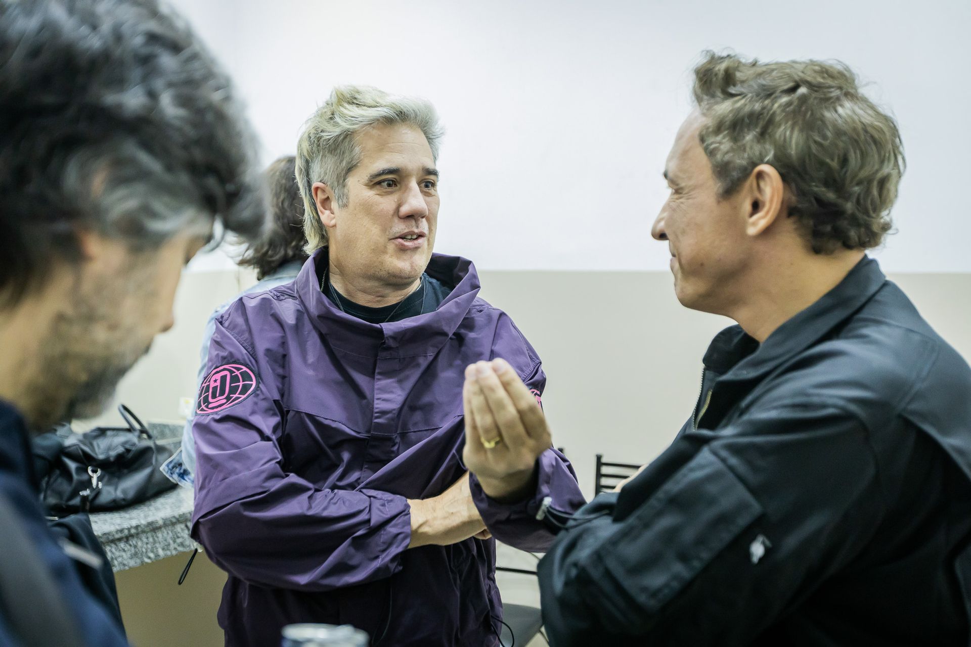 Duas pessoas conversavam, uma vestindo uma jaqueta roxa e gesticulando com a mão, a outra vestindo uma jaqueta preta.