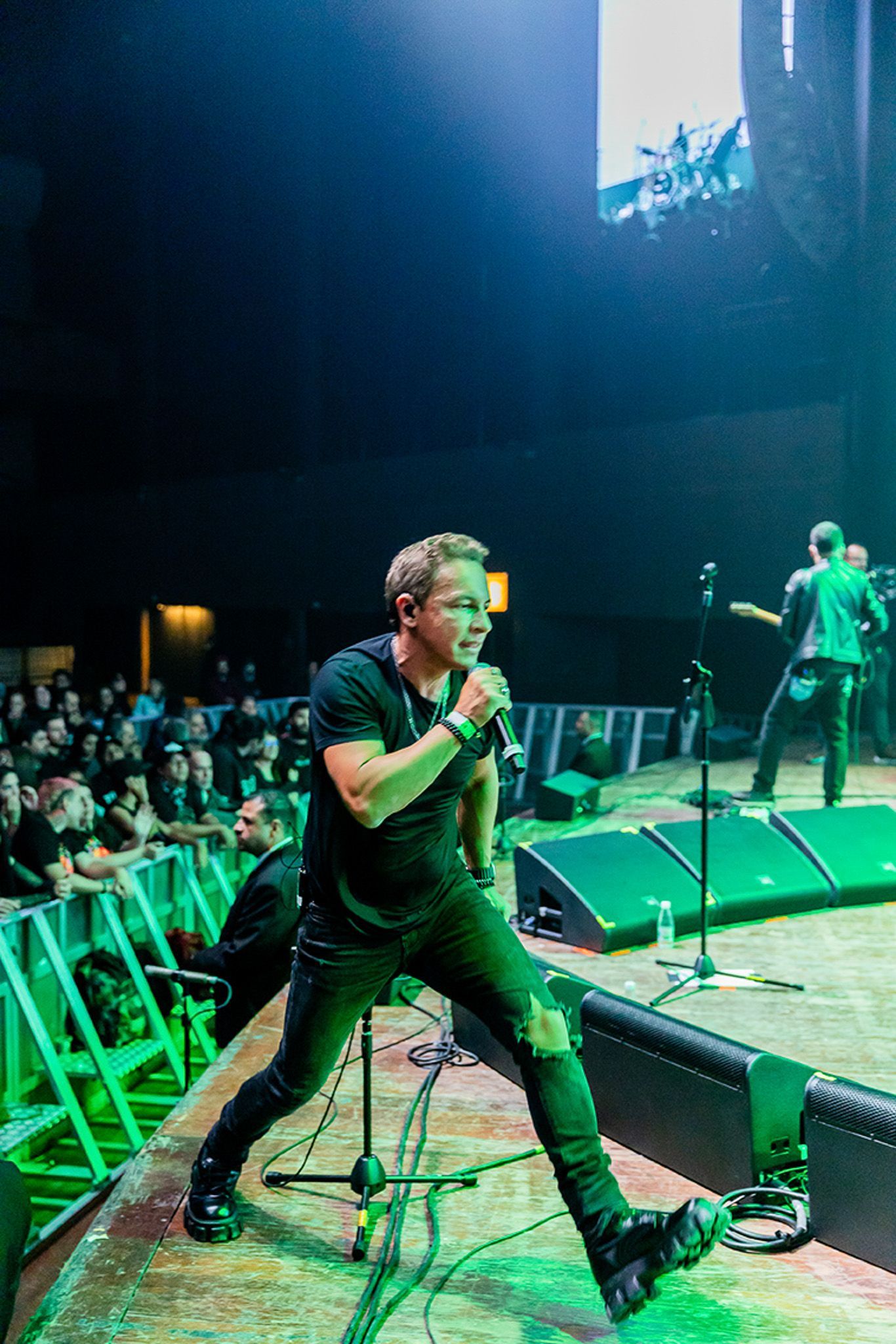 Um artista cantando em um microfone em um palco de concerto, com um músico ao fundo e uma plateia em primeiro plano.