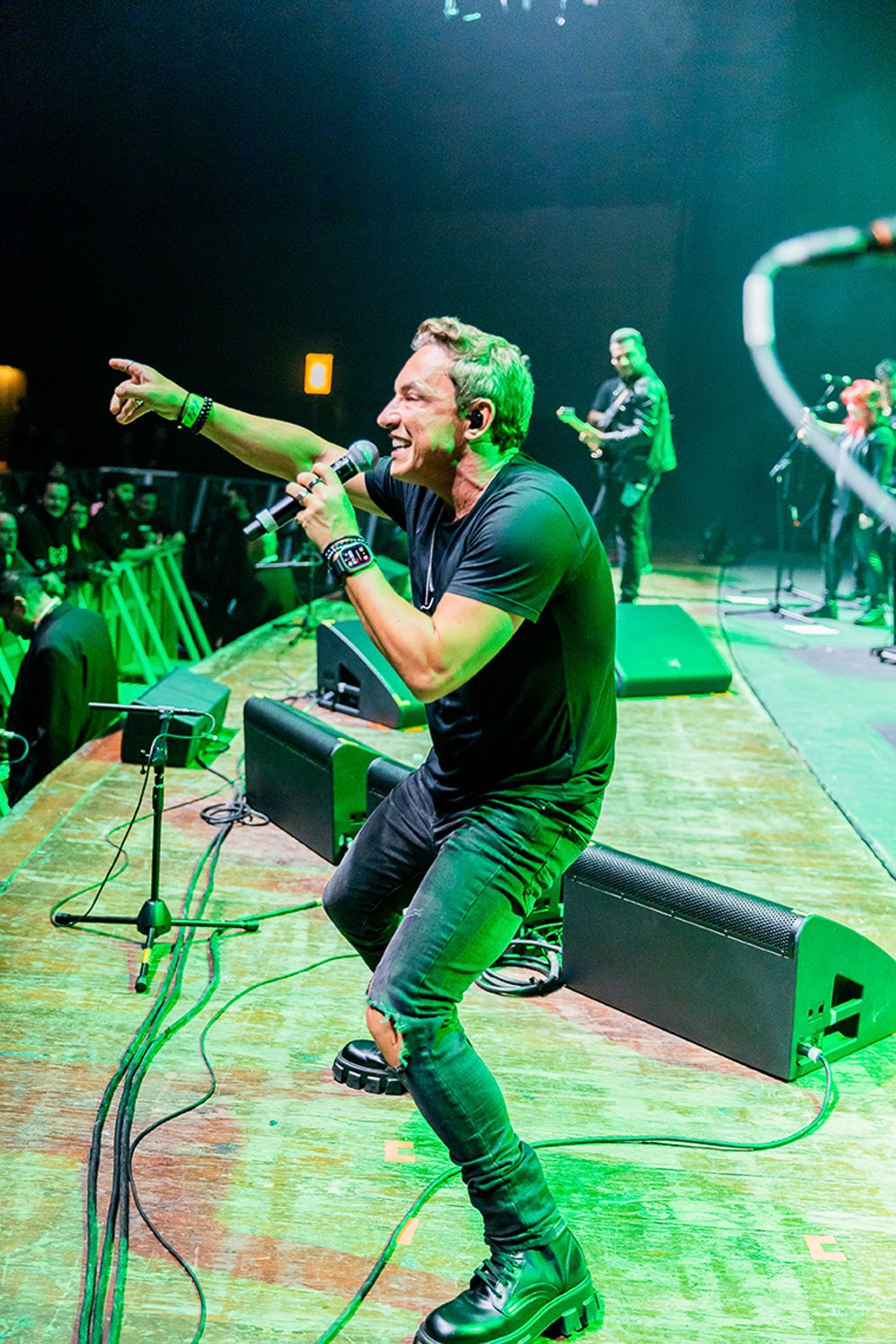 Um artista vestindo camisa preta e calça jeans segura um microfone e aponta para a plateia enquanto está em um palco.