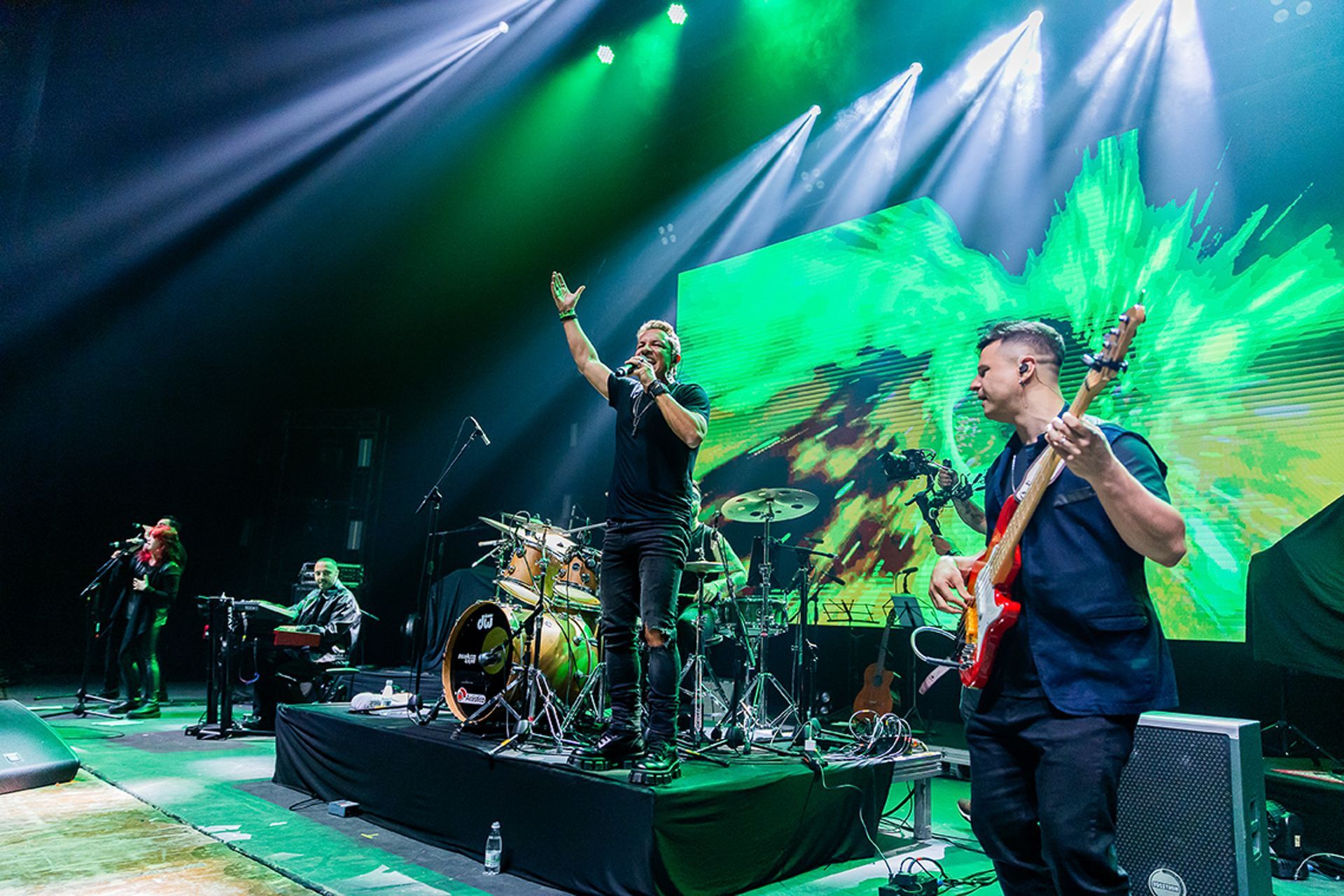 Uma banda se apresentando no palco com luzes verdes, um vocalista com as mãos levantadas e um guitarrista.