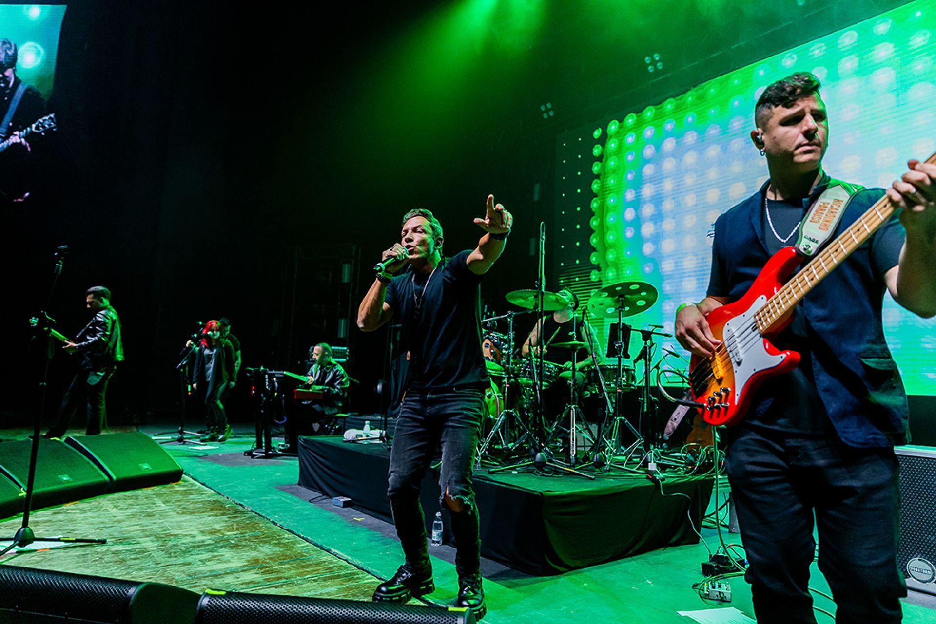 Uma banda se apresenta em um palco iluminado com luzes verdes; um vocalista canta enquanto um baixista toca uma guitarra.
