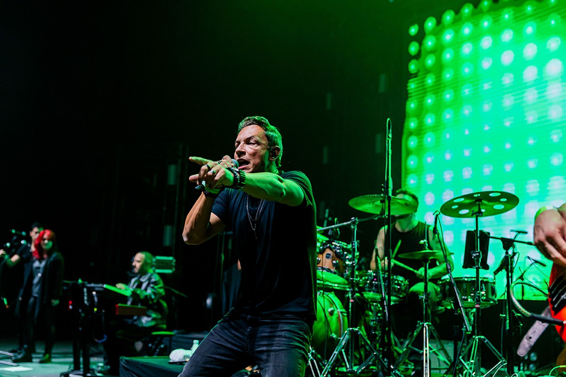 O vocalista principal se apresenta no palco com uma banda sob luzes verdes, gesticulando com as mãos em direção à plateia.