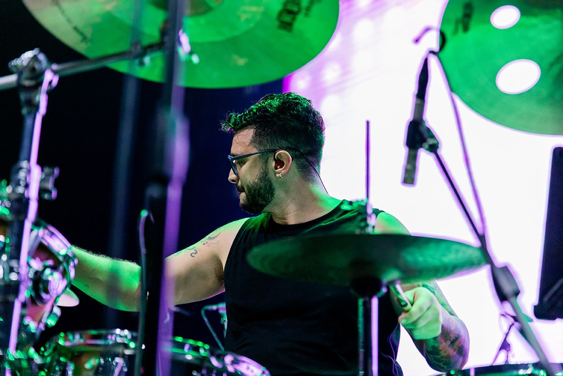 Um baterista usando óculos e uma regata preta toca em um palco iluminado por luzes de palco verde-brilhantes.