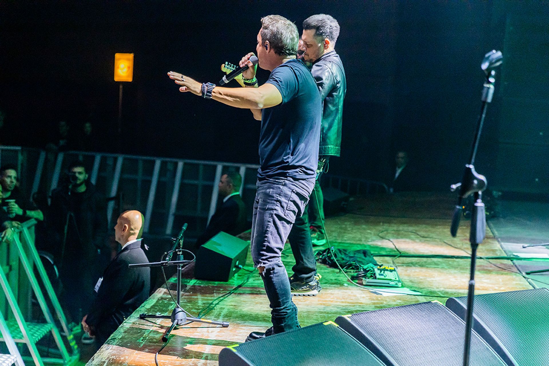 Dois artistas vestidos de preto em um palco; um gesticula para a plateia enquanto segura um microfone