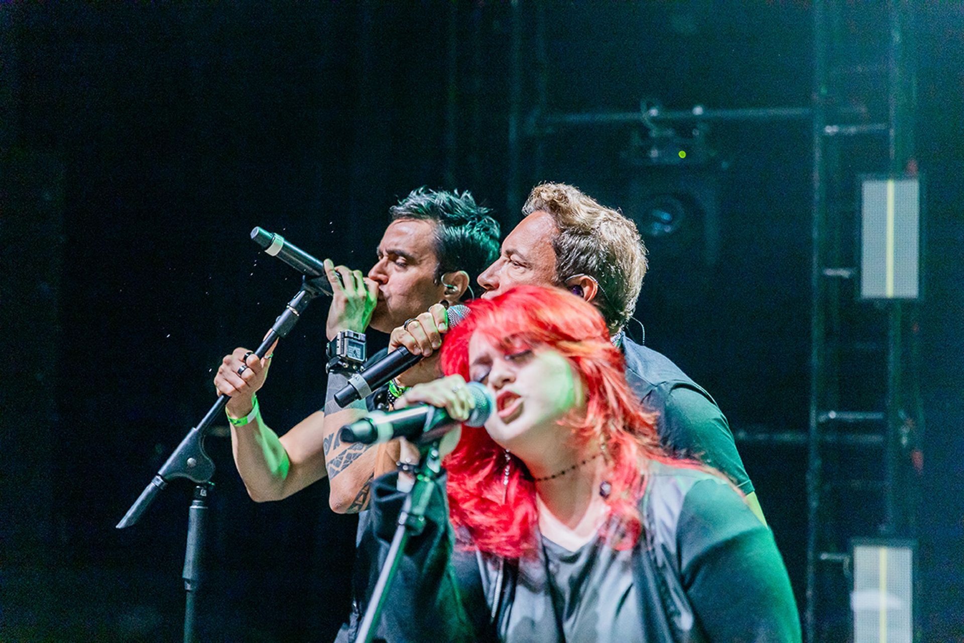 Uma mulher com vibrantes cabelos ruivos e dois homens cantando em microfones em um palco com pouca luz.