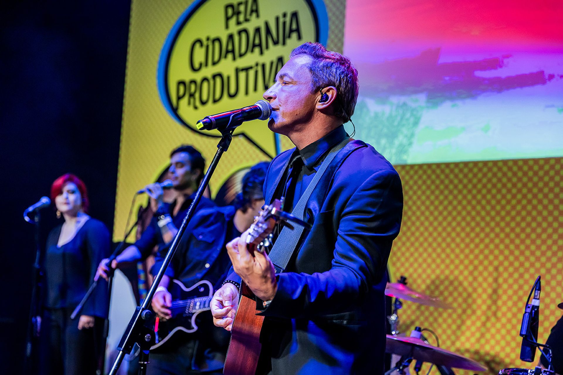 Uma banda se apresenta no palco. Uma faixa amarela com os dizeres 