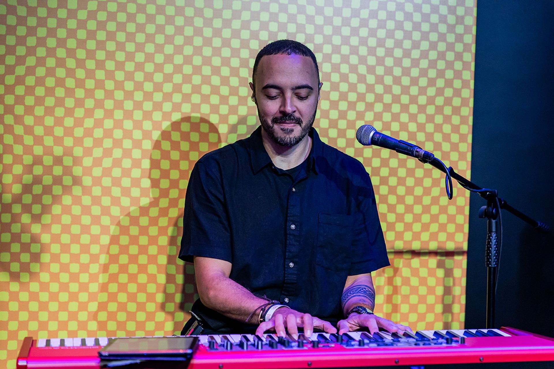 Um músico tocando teclado com um microfone em frente a um fundo quadriculado amarelo.