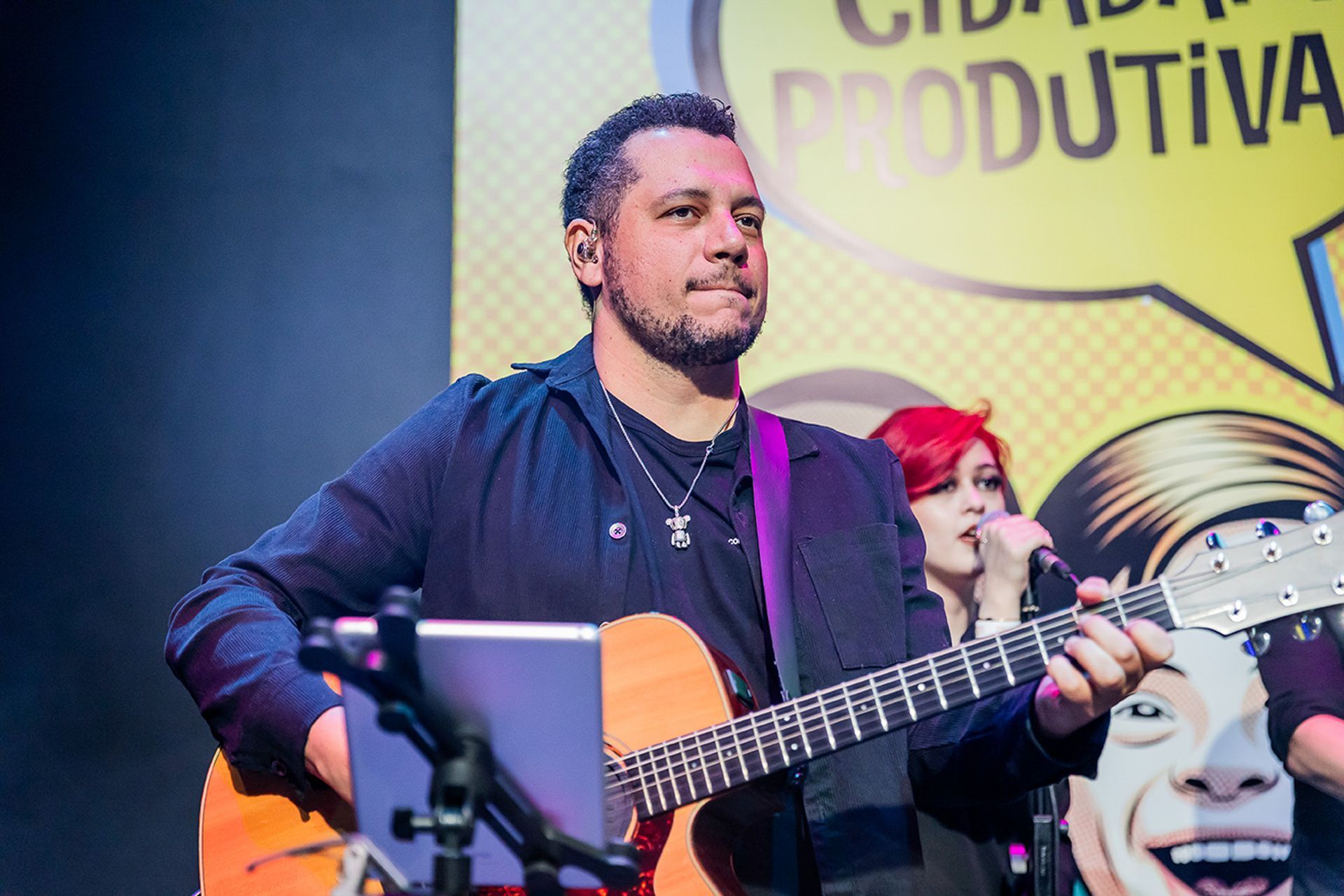 Um artista toca violão no palco em frente a uma faixa amarela com os dizeres 