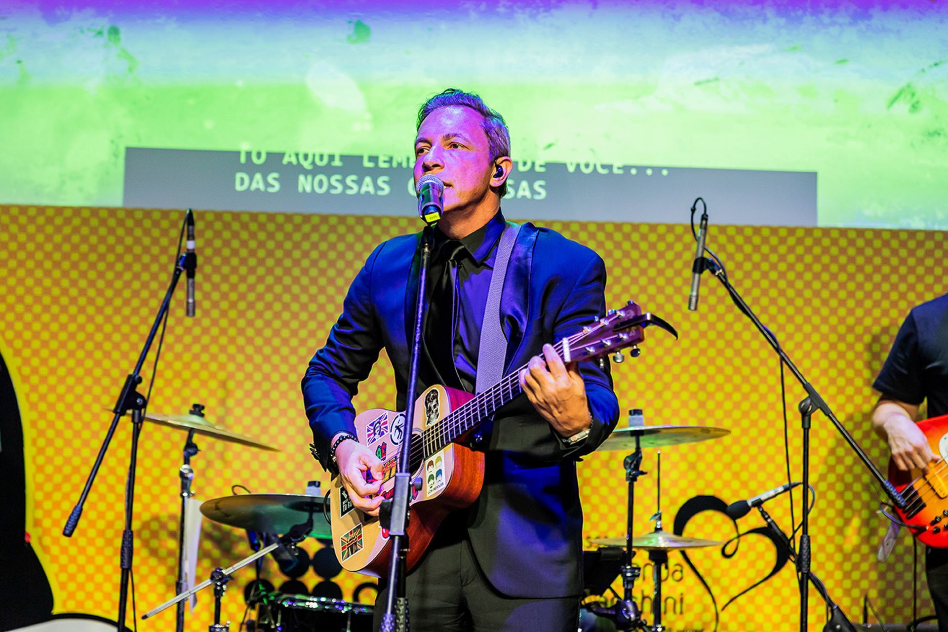 Um músico de terno toca violão e canta ao microfone em um palco com painéis de fundo amarelos.