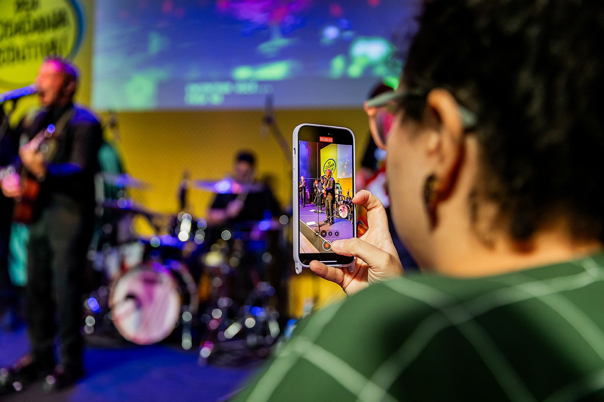Uma pessoa em primeiro plano filma uma banda se apresentando ao vivo no palco com um smartphone.