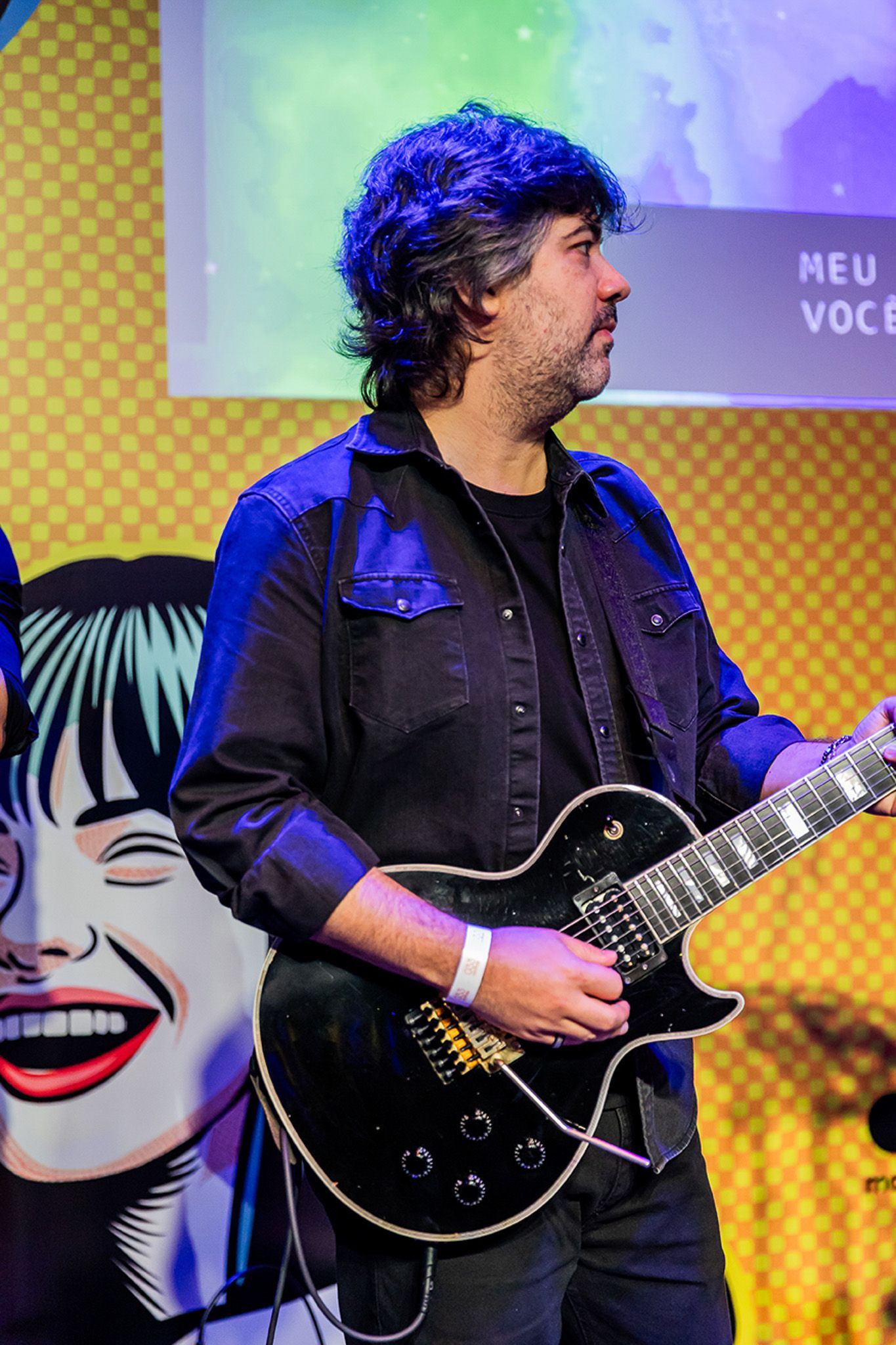 Um guitarrista de cabelos escuros e toca uma guitarra, vestindo uma camisa escura de botões. fundo amarelo de arte pop.