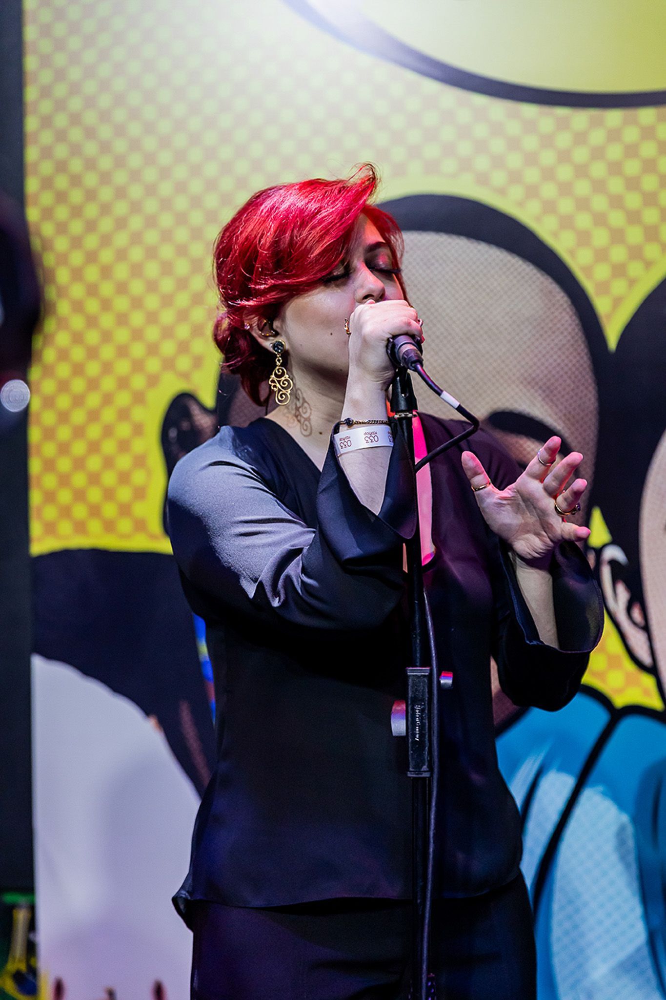 Uma pessoa com cabelos ruivos vibrantes cantando em um microfone no palco, em frente a um cenário colorido e estampado.