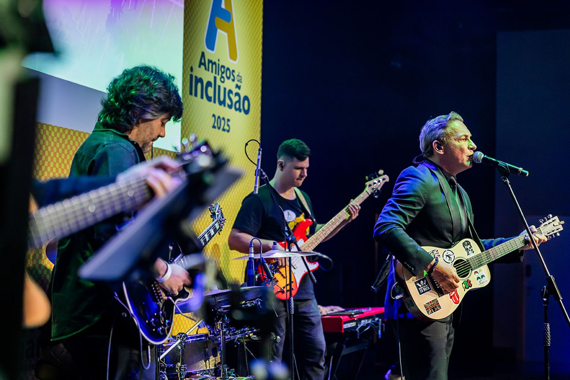 Três músicos se apresentam em um palco com uma faixa que diz 