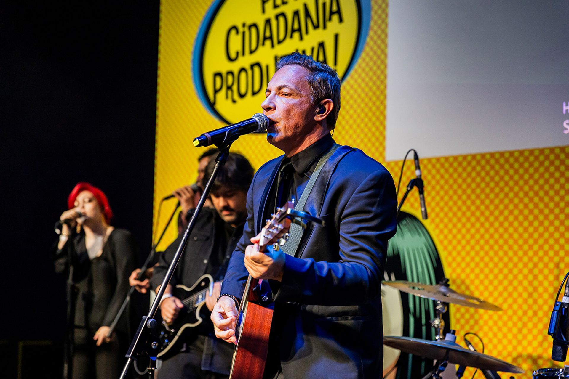Um músico toca violão e canta em um microfone no palco, com uma banda e um cenário amarelo atrás dele.