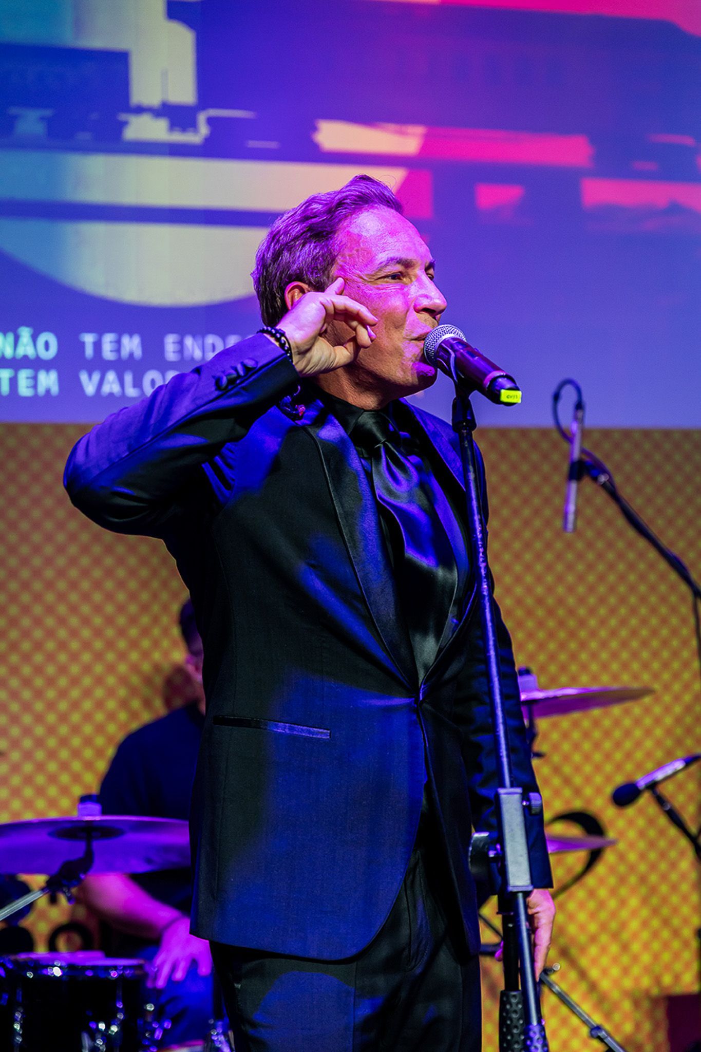 Um artista de terno preto cantando em um microfone no palco, com um fundo claro e instrumentos musicais visíveis.