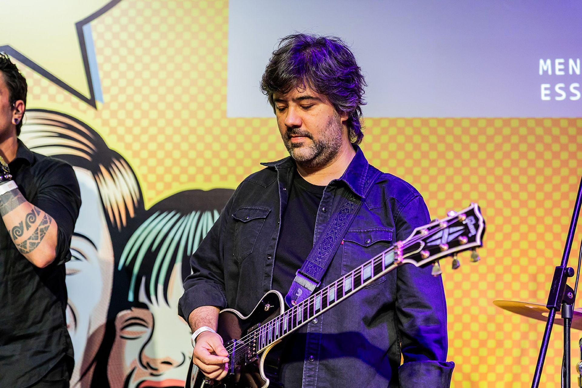 Um músico barbudo tocando guitarra elétrica no palco em frente a um cenário colorido e estampado.