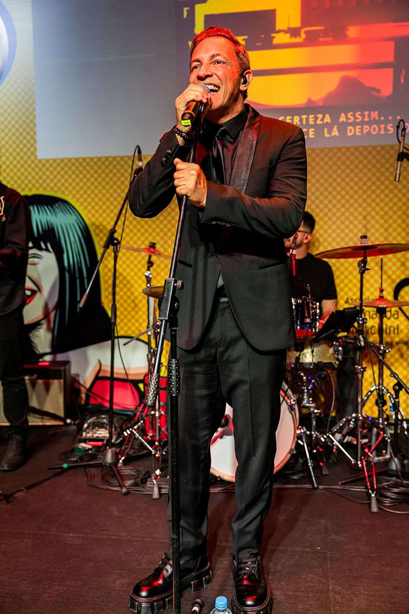 Um cantor de terno preto se apresenta no palco, segurando um microfone em frente a um fundo amarelo com um mural de arte