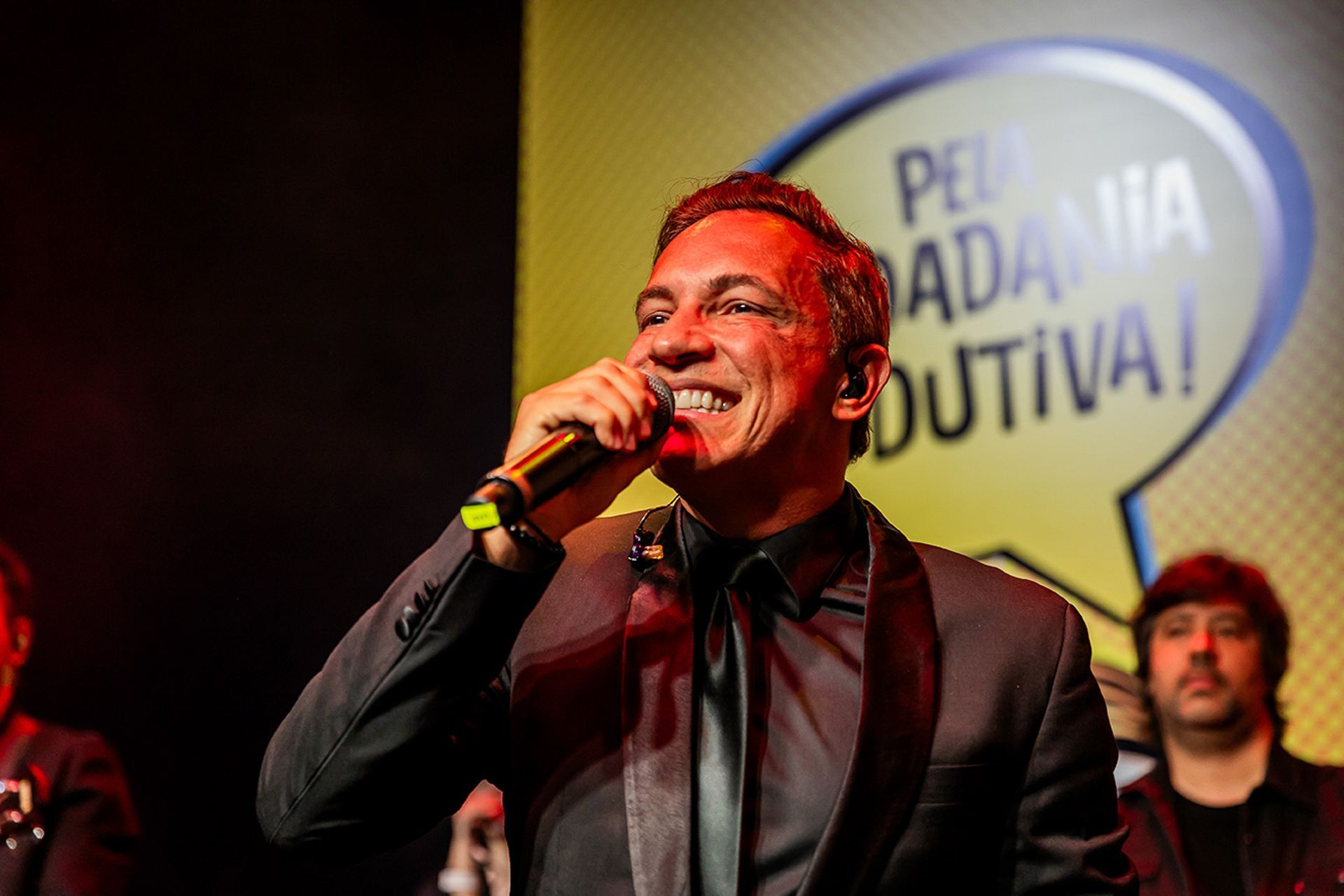 Um homem de terno e gravata pretos canta em um microfone no palco, em frente a um fundo de balões de fala amarelos.