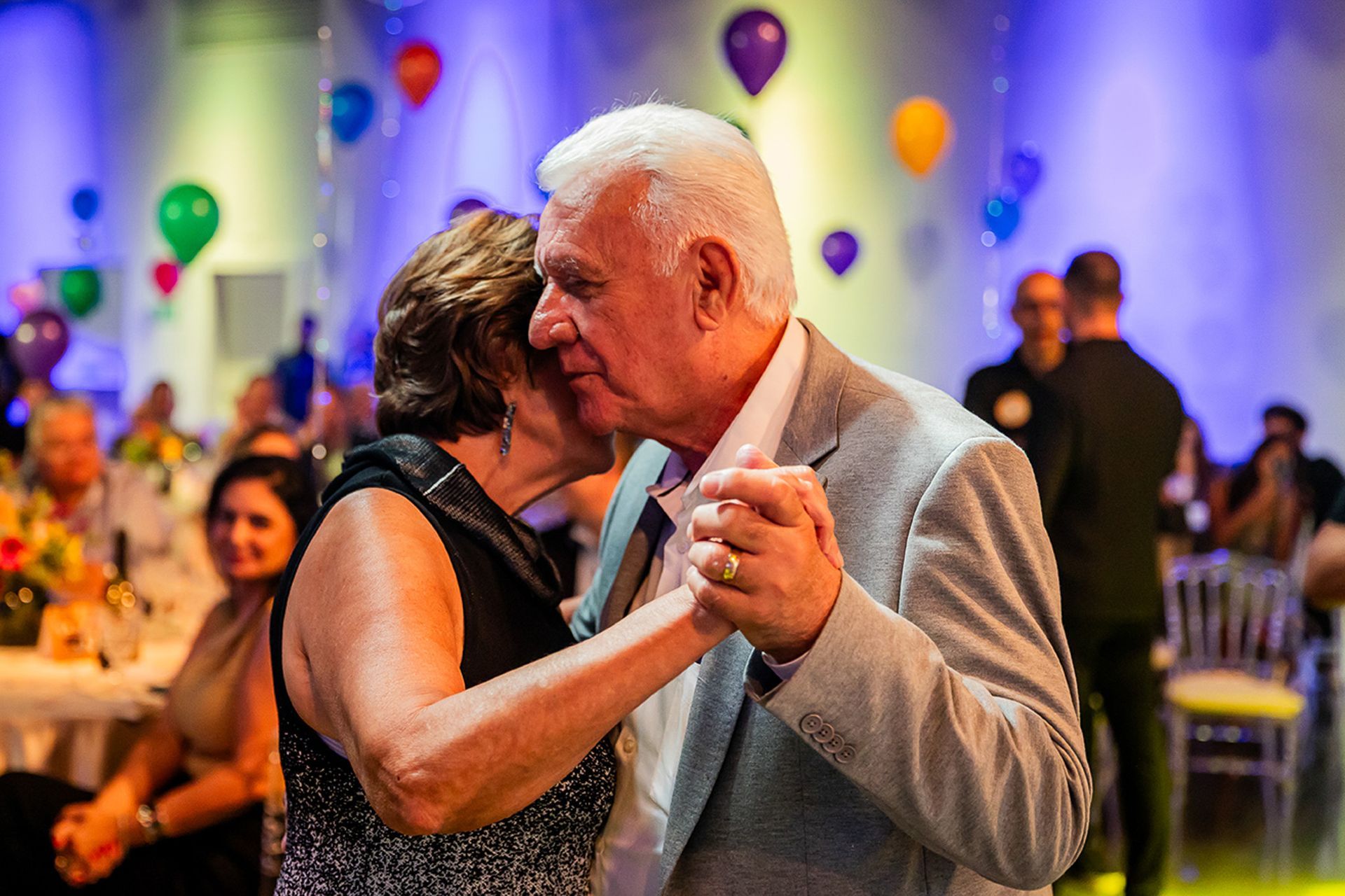 Duas pessoas dão as mãos e se abraçam enquanto dançam em uma festa decorada com balões coloridos.